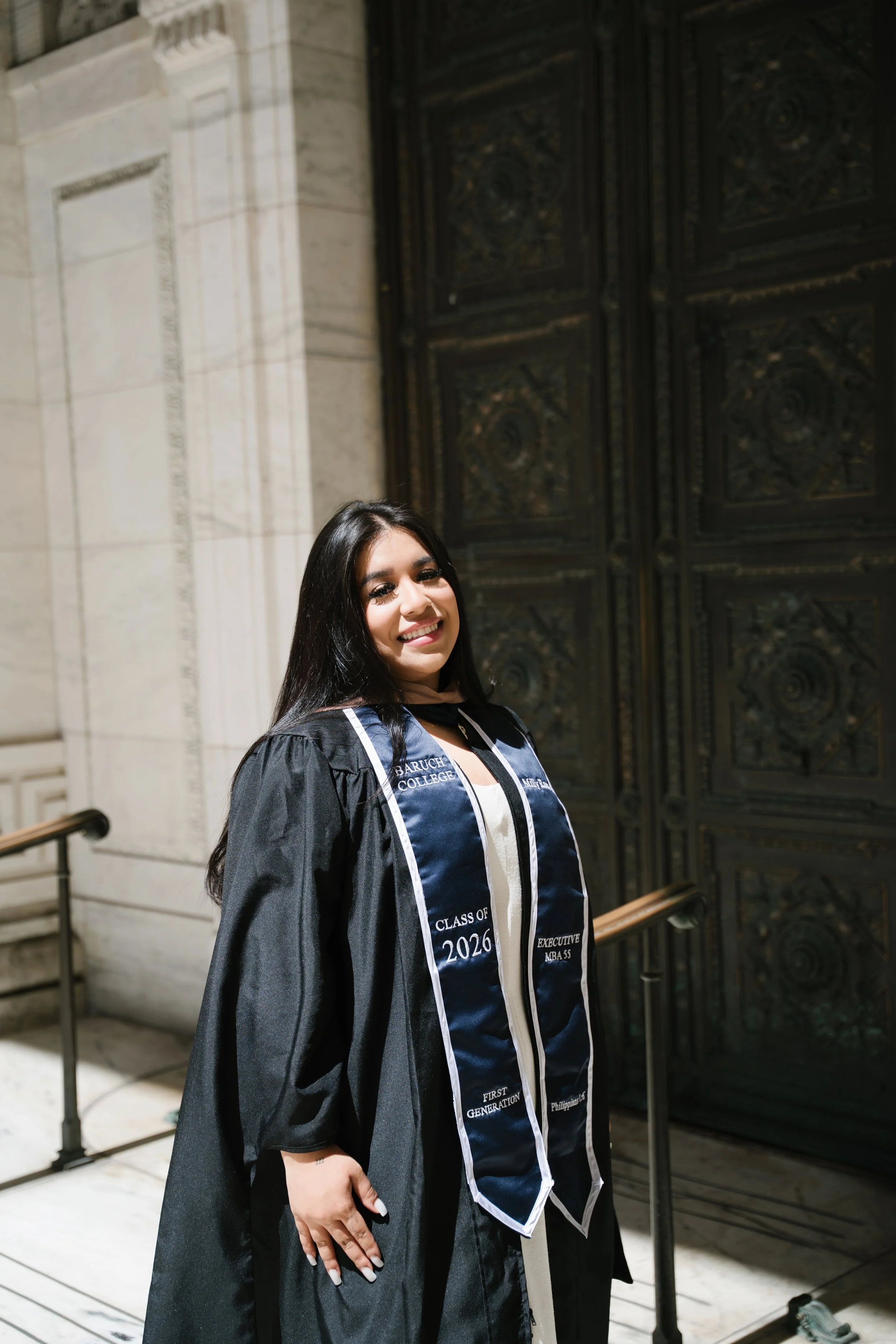 Young woman in graduation gown and sash standing in front of a large ornate door in a historical building, smiling at the camera.