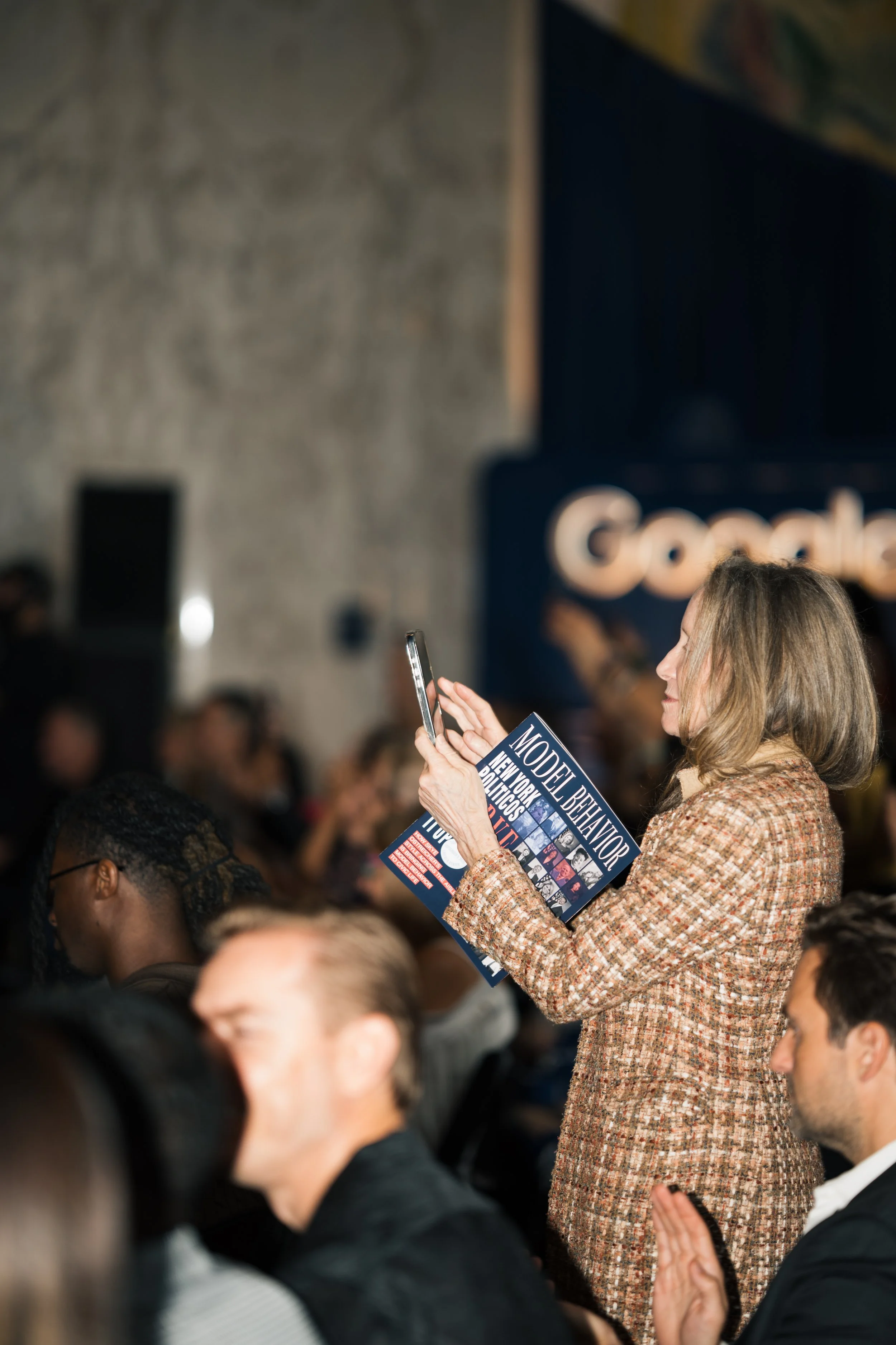 A woman with long blonde hair wearing a brown and beige tweed coat holding a magazine and a smartphone at a crowded event, with people seated and a dark background featuring a sign.