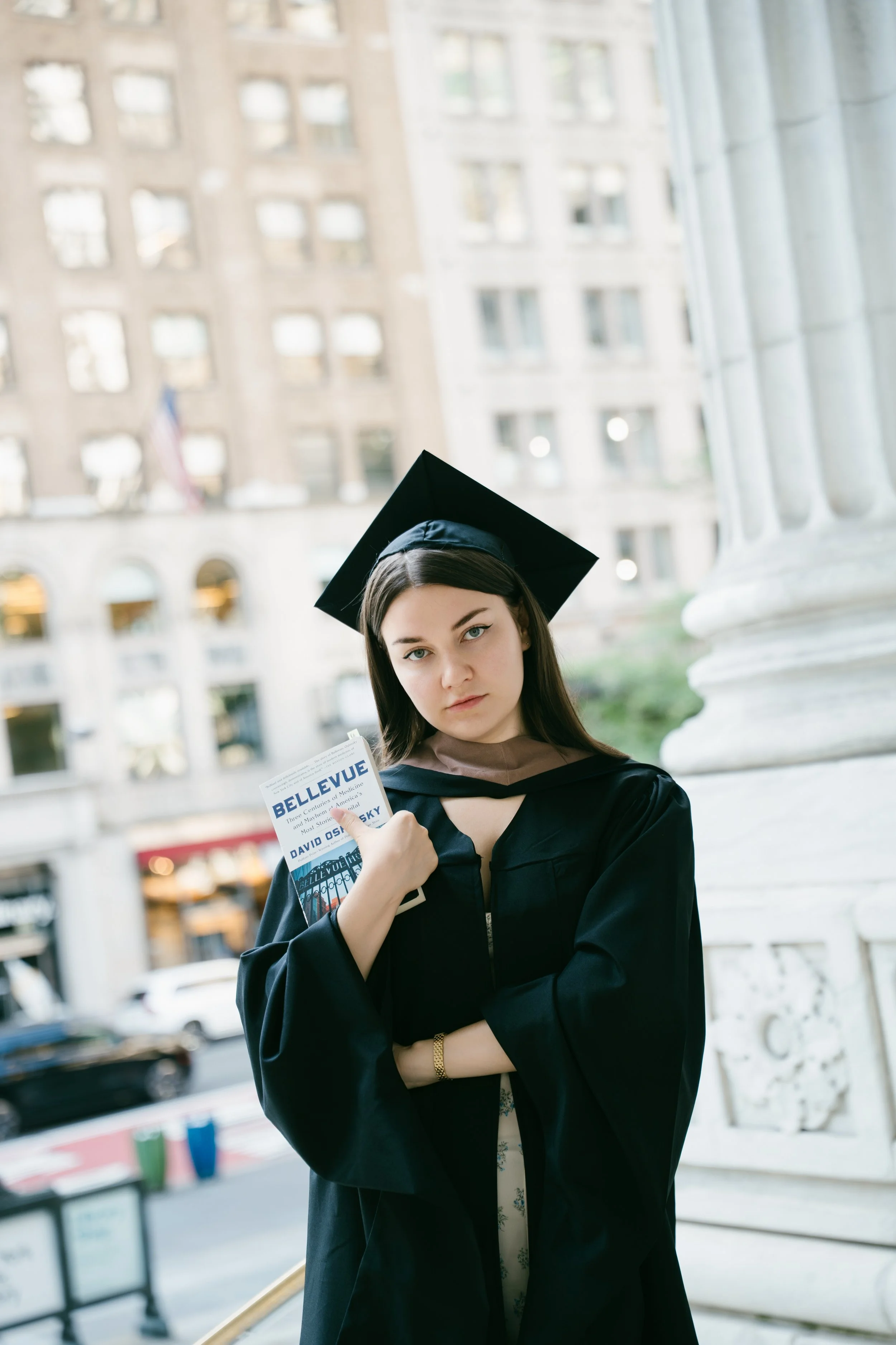 Young woman in a graduation cap and gown holding a book titled "BELLEVUE", standing outdoors in front of a large column with city buildings in the background.
