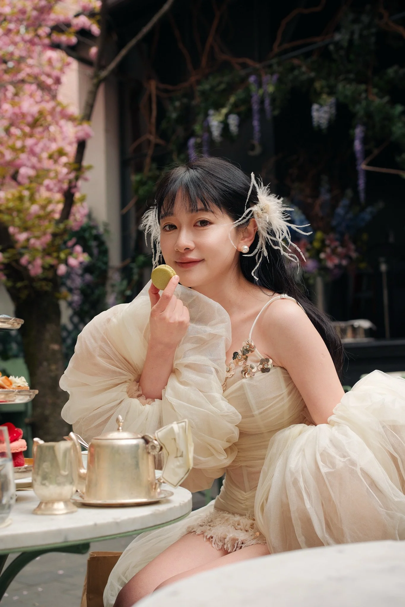 A woman dressed in an elegant, ivory, ruffled gown with floral embellishments, sitting at a table set for tea. She has dark hair adorned with decorative feathers and pearl earrings. She is holding a green macaron to her lips and smiling slightly. The