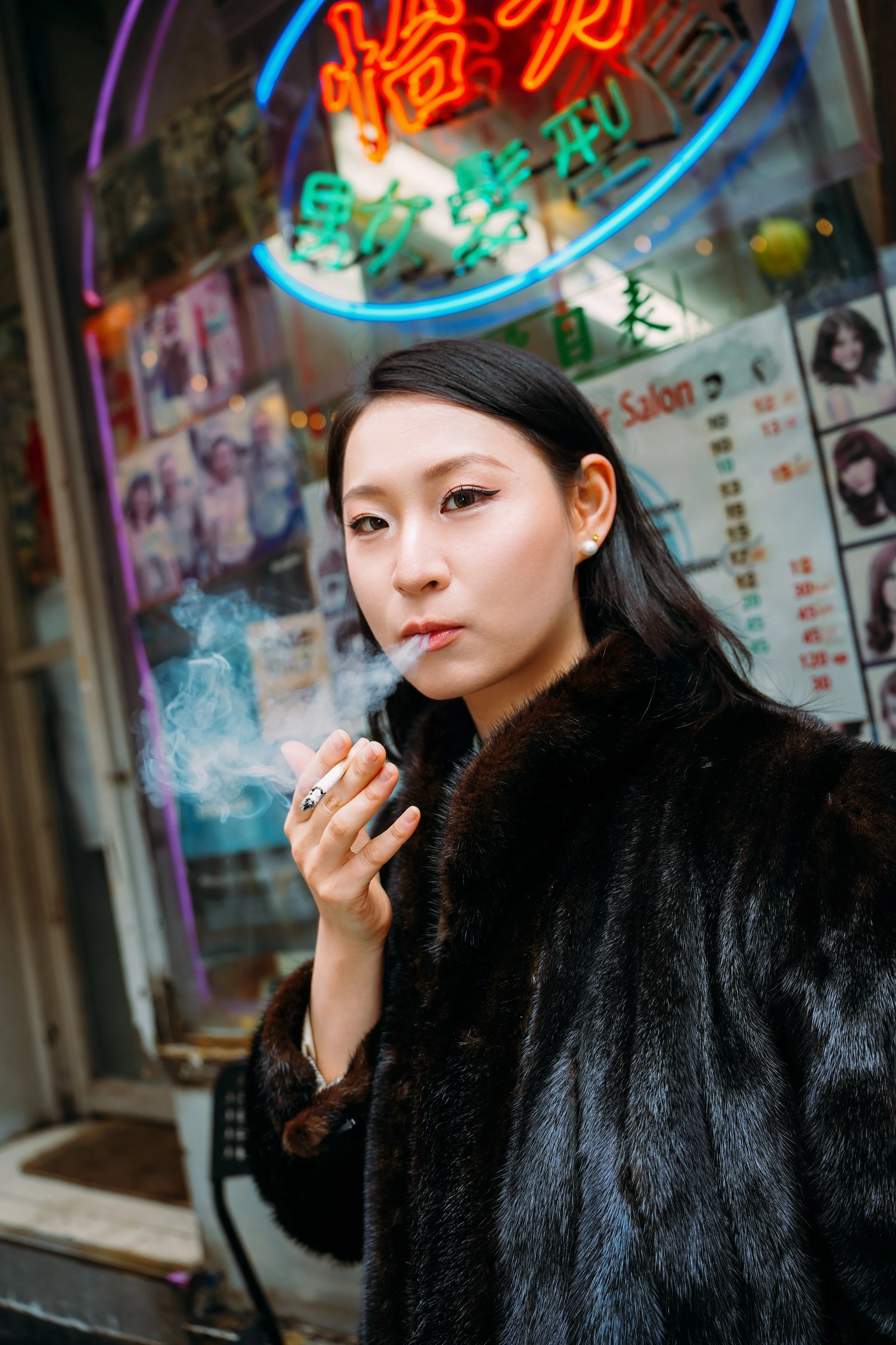 A young woman with dark hair, wearing a fur coat and pearl earrings, stands outside a store with colorful neon signage, smoking a cigarette with smoke visible.