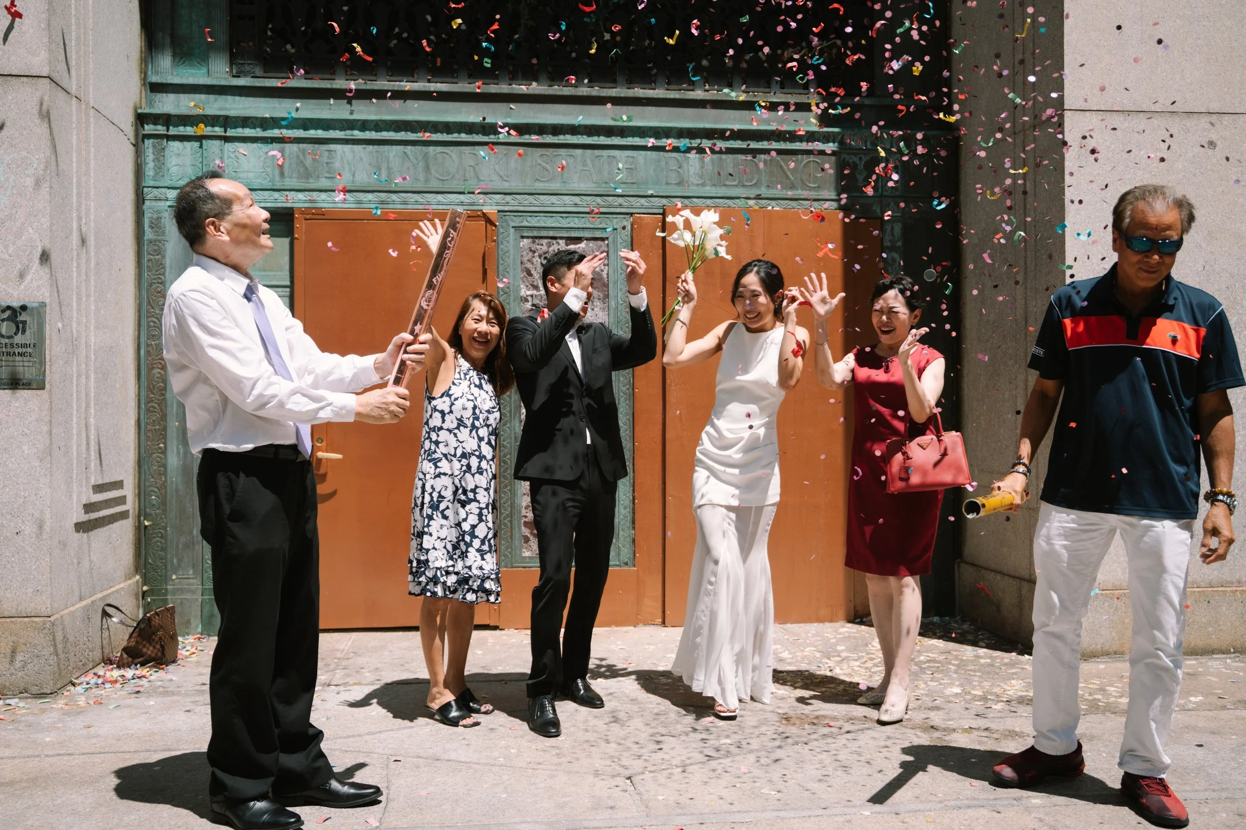 People celebrating outside in front of a building with confetti falling and a person holding flowers.