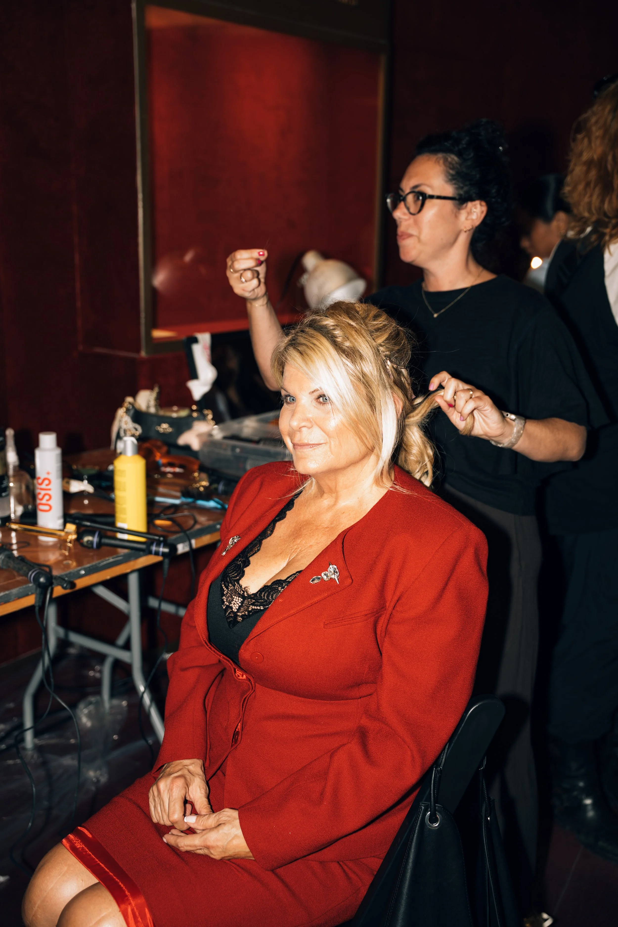 A woman with blonde hair wearing a red blazer and lace top sitting in a salon chair, getting her hair styled by a stylist behind her. The stylist is a woman in black shirt and glasses, holding a section of hair. The salon has a dark wood wall and sev