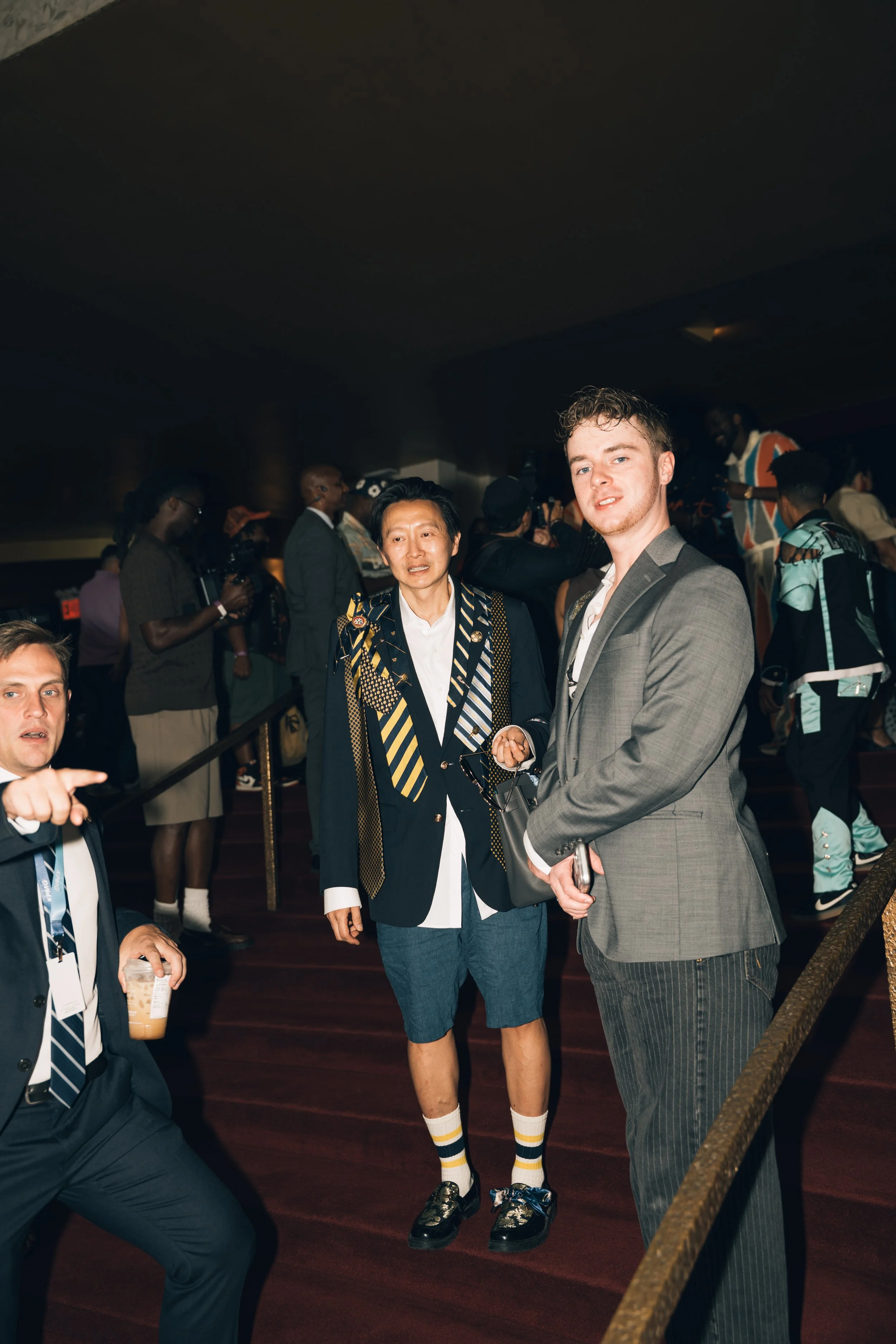 A group of people socializing at an indoor event. The focus is on two men standing in the foreground, one in a suit and the other in a blazer, shorts, and sneakers. Other attendees are visible in the background, with some holding cameras or drinks.