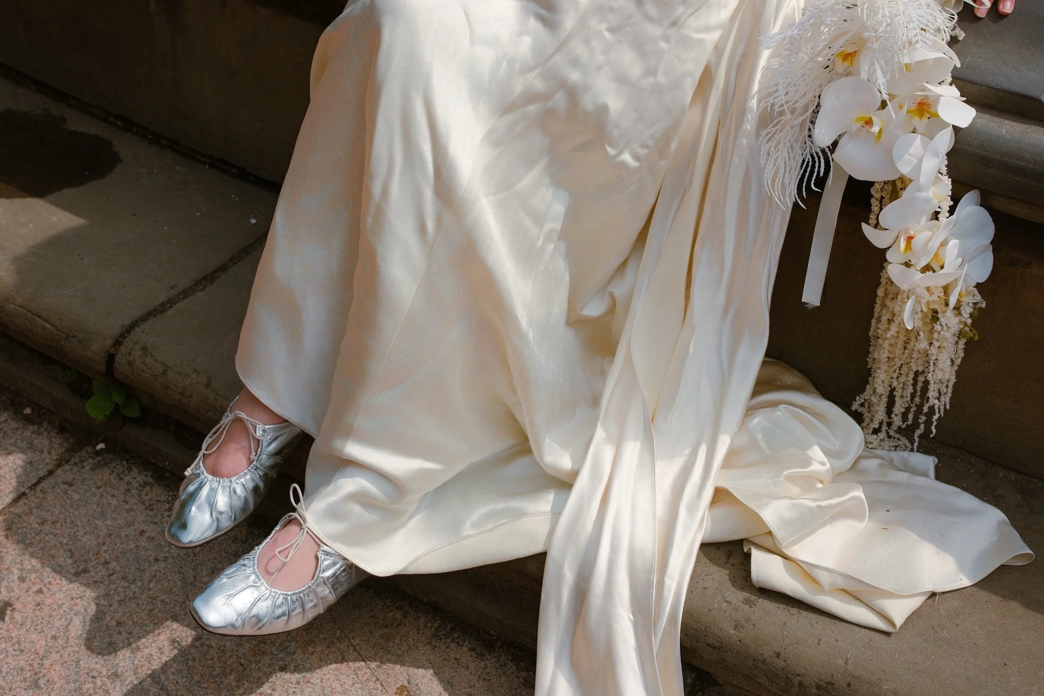 A person in a cream-colored dress sits on the ground with metallic silver ballet flats. They are next to a floral arrangement with white orchids and hanging white flowers, placed on a bench against a stone surface.