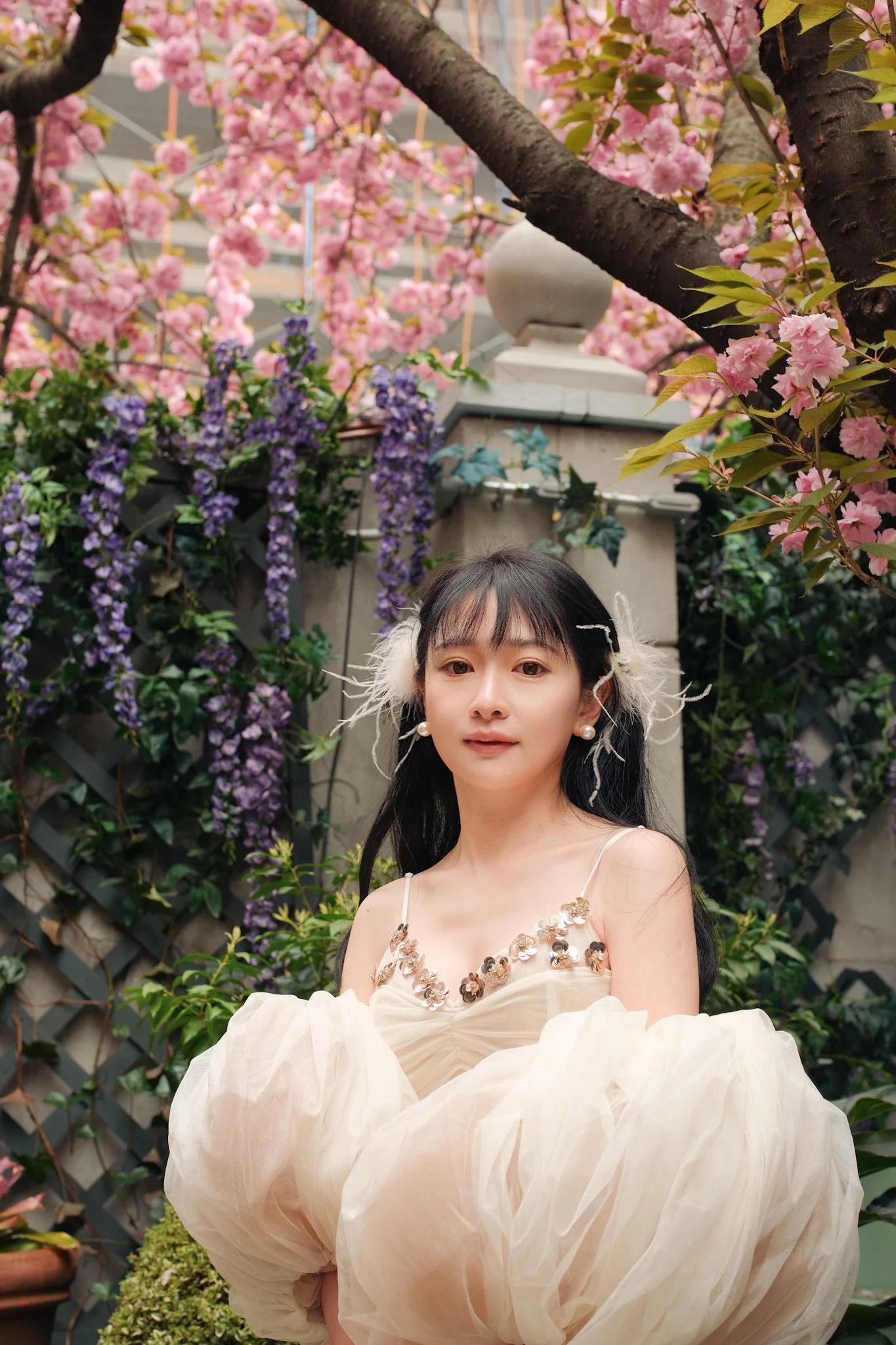 A young woman with black hair and pearl earrings taking a selfie amidst pink cherry blossoms and purple wisteria flowers in a garden.