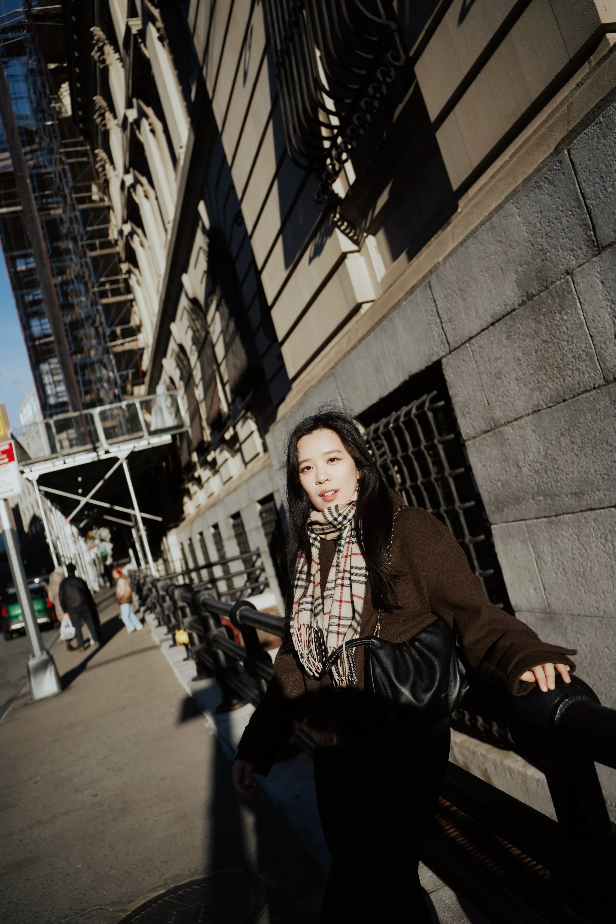 A young woman with long dark hair wearing a brown coat and Burberry scarf, sitting on a city bench with a stone building and scaffolding in the background. She is looking at the camera with a neutral expression.