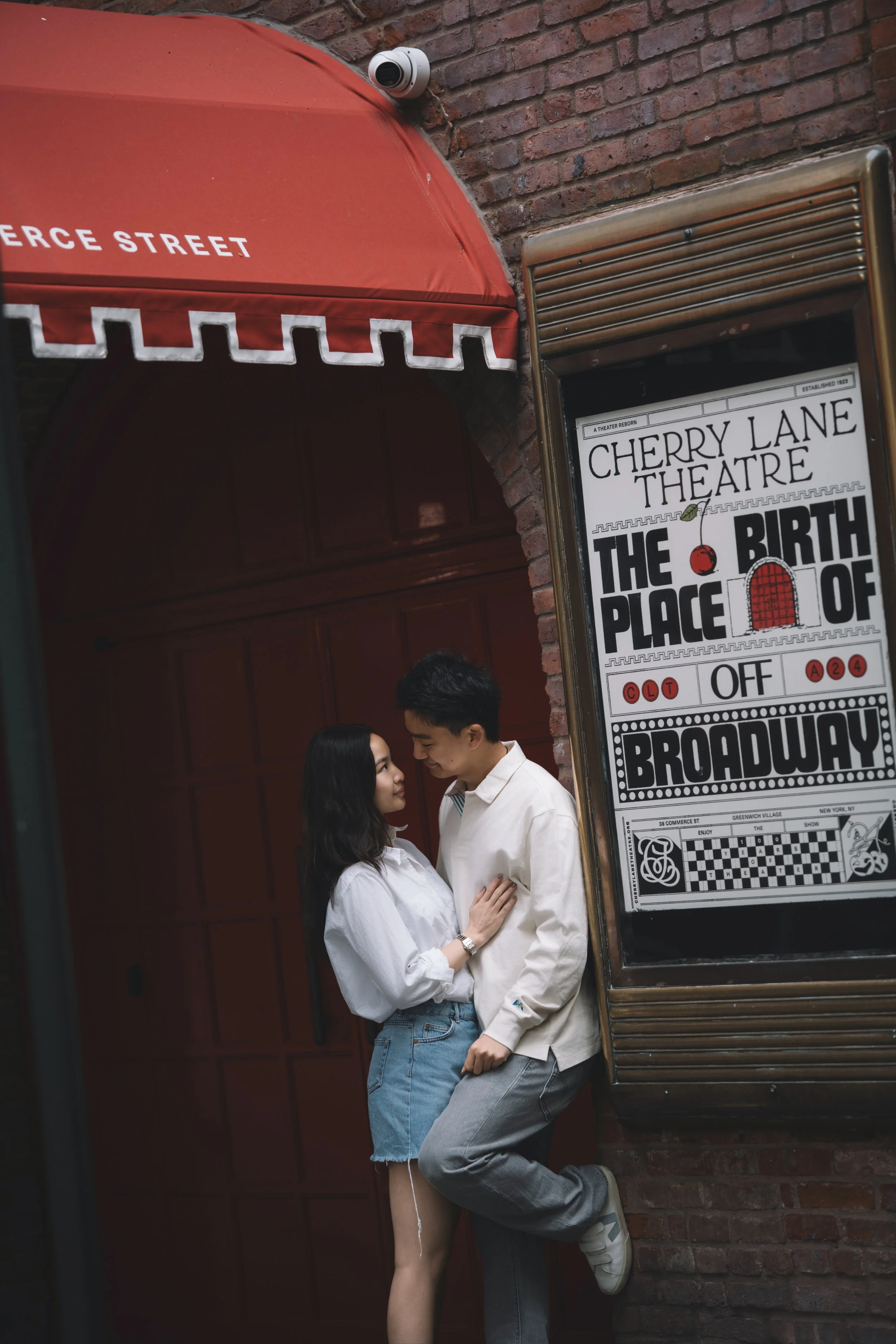 A young couple stands close together outside a theater on Cherry Lane, with the woman touching the man's chest, both smiling and gazing into each other's eyes. The theater's sign and brick wall are visible in the background.