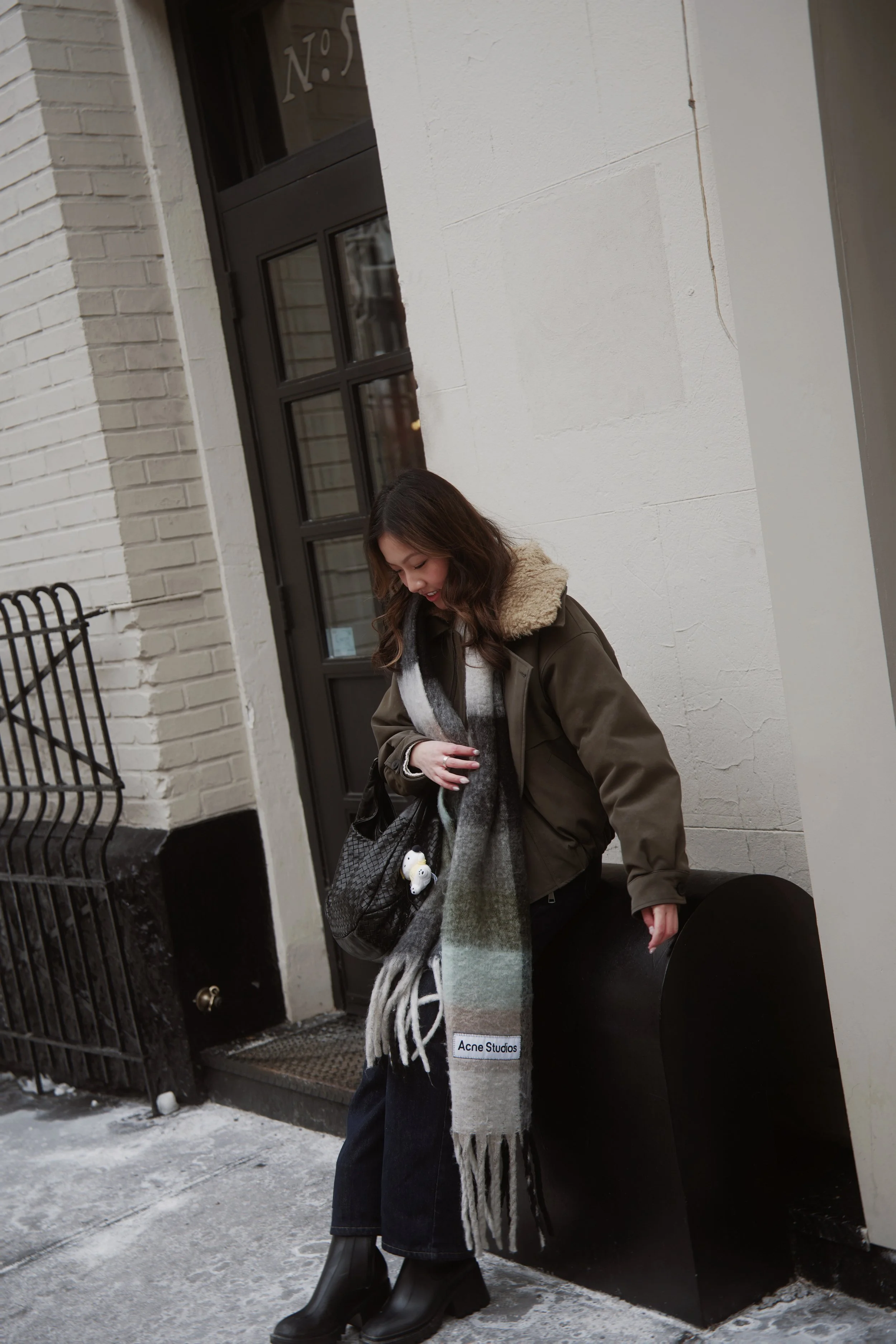 A young woman with wavy brown hair wearing a green jacket with a beige fur collar, a gray plaid scarf, black pants, and black boots, standing outside on a snowy sidewalk next to a black mailbox and a white brick wall.