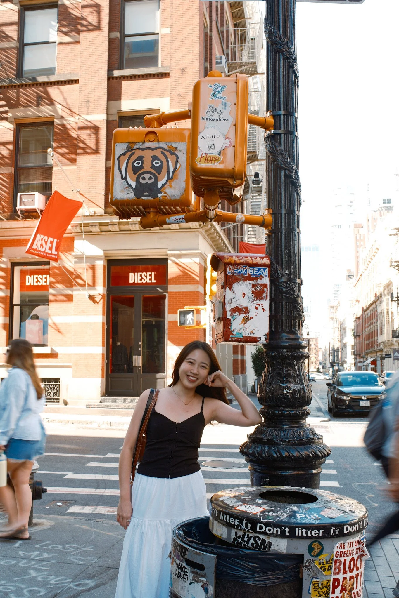 A young woman with long dark hair smiling and making a heart shape with her hand near her face, standing on a city street corner beside a black trash can covered in stickers and graffiti. Behind her are pedestrians, brick buildings, and parked cars.