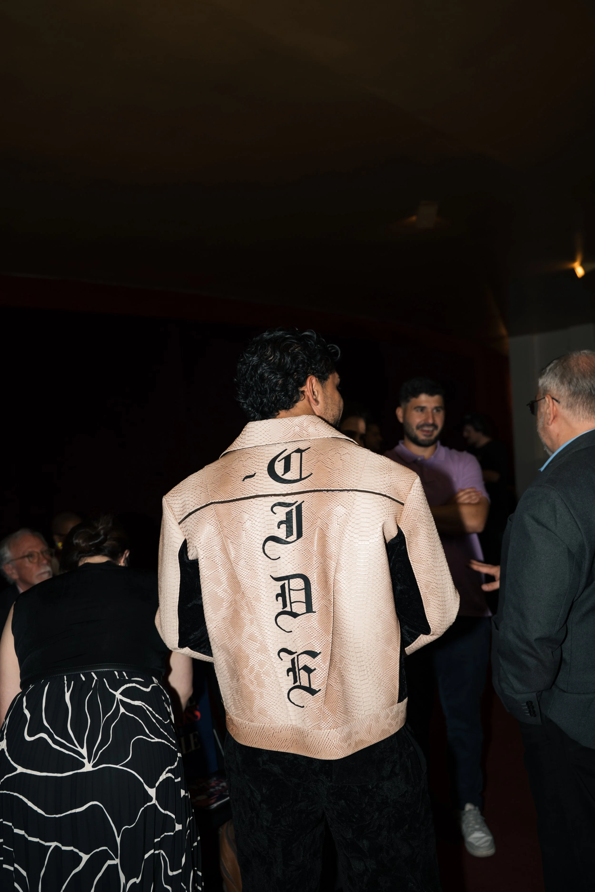 A man with curly hair wearing a beige jacket with black gothic lettering on the back is standing and talking with an older man in a suit. There are other people in the background, including a man in a purple shirt and a woman with dark hair and glass
