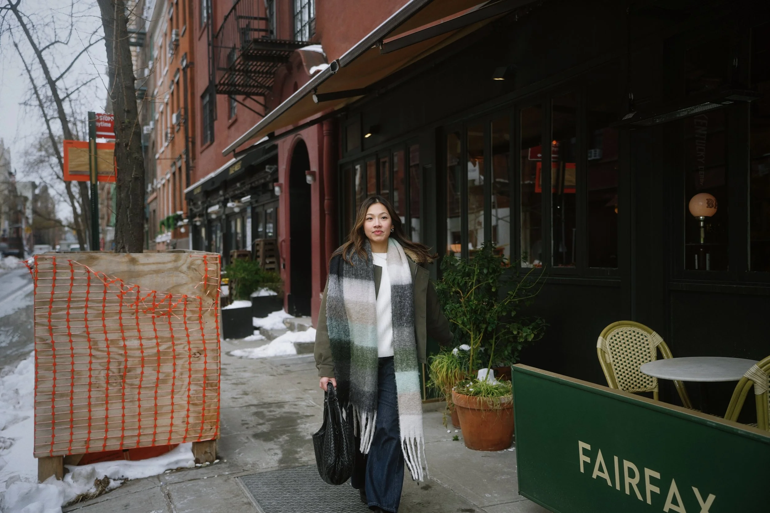A woman walking on a city sidewalk on a cold day, wearing a large scarf and coat, with a bag in her hand. There are brick buildings, leafless trees, and a green vegetable cart with a sign that reads 'Fairway' nearby.