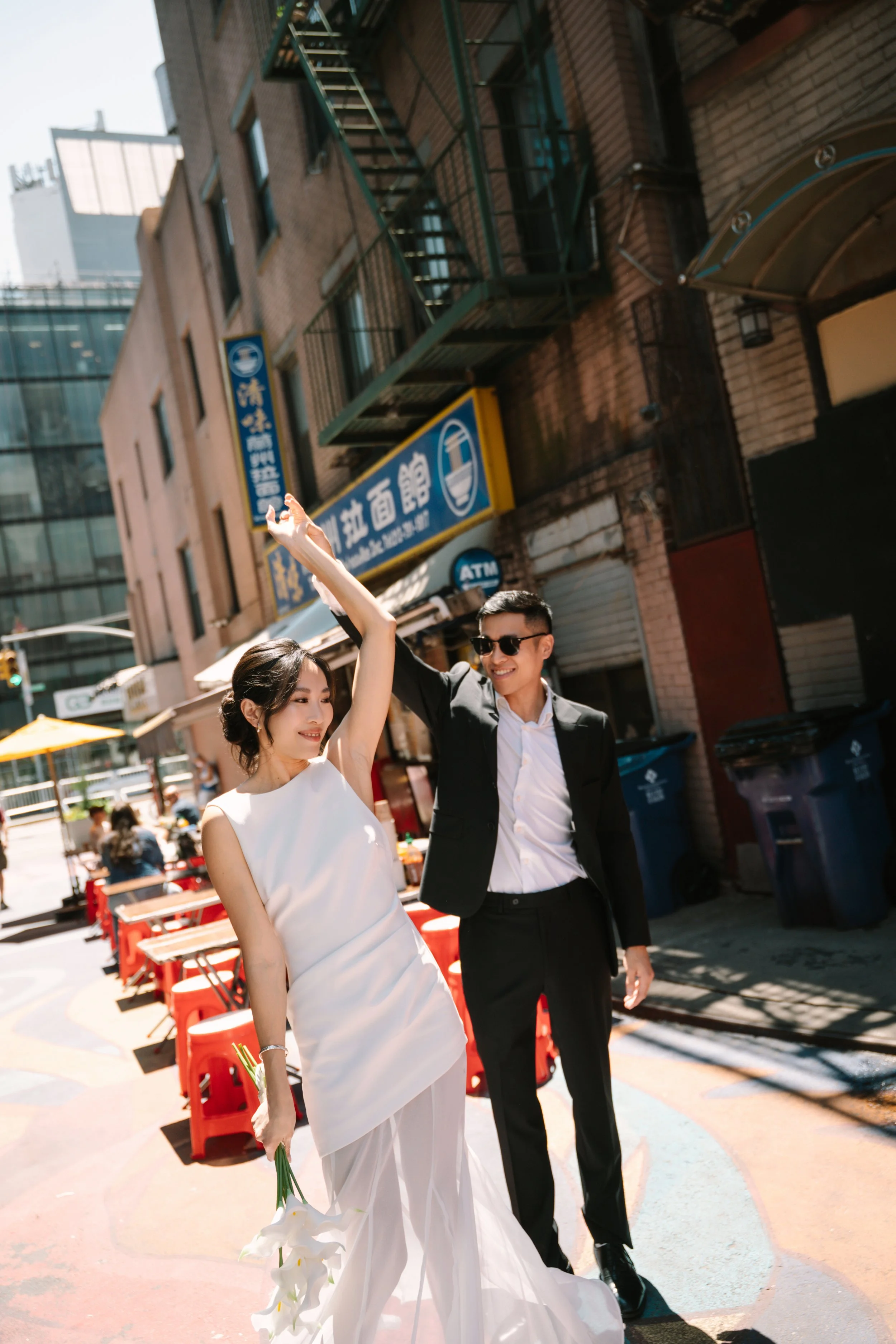 A bride in a white wedding dress holding a bouquet of white lilies, smiling, with a groom in a black suit and sunglasses, on a busy city street with red outdoor chairs, yellow umbrellas, and storefront signs in Asian characters.
