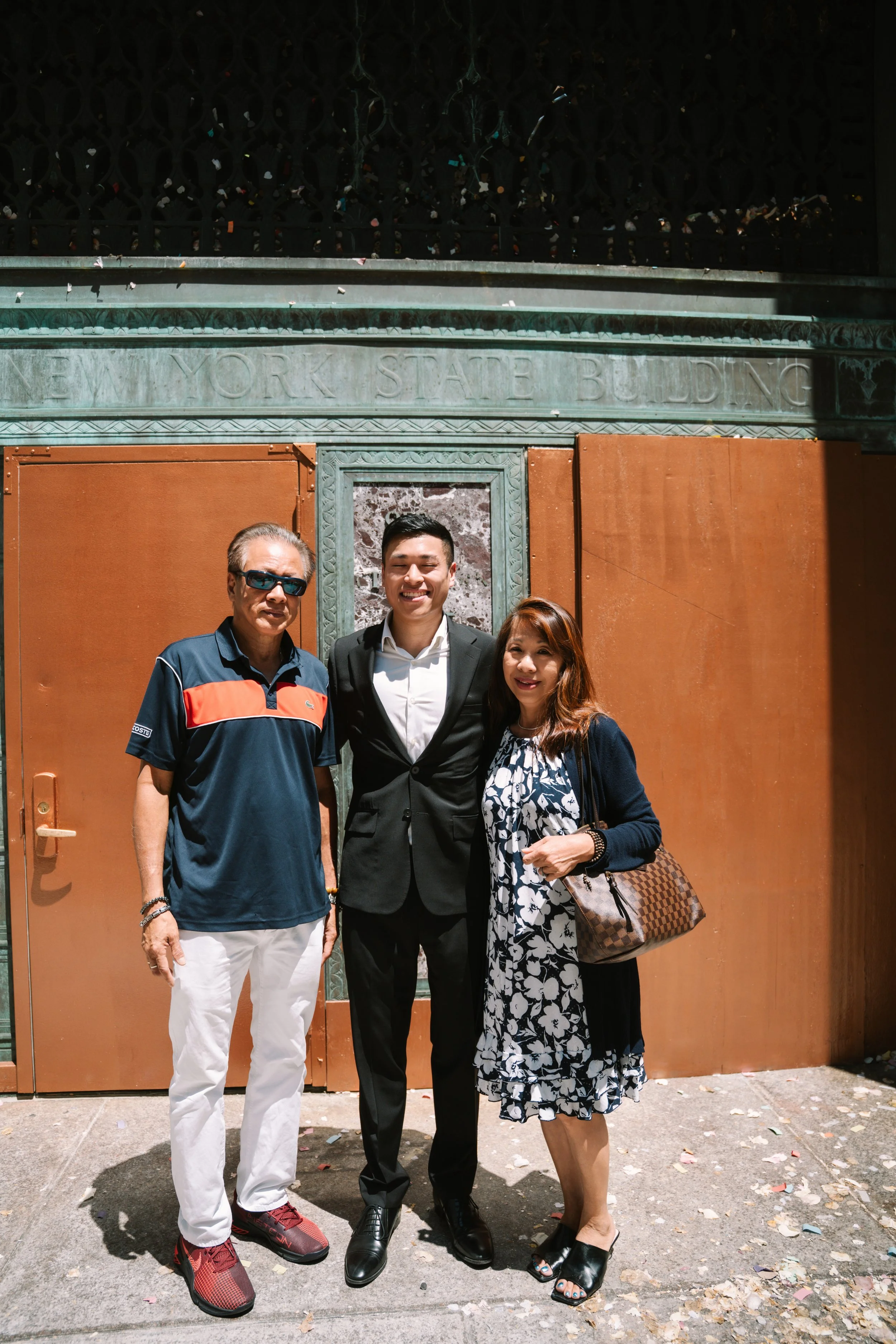 Three people standing in front of a building with the words 'YORK STATE BUILDING' engraved on it. The man on the left wears sunglasses, a navy polo with red and white stripes, white pants, and red sneakers. The man in the middle wears a black suit wi