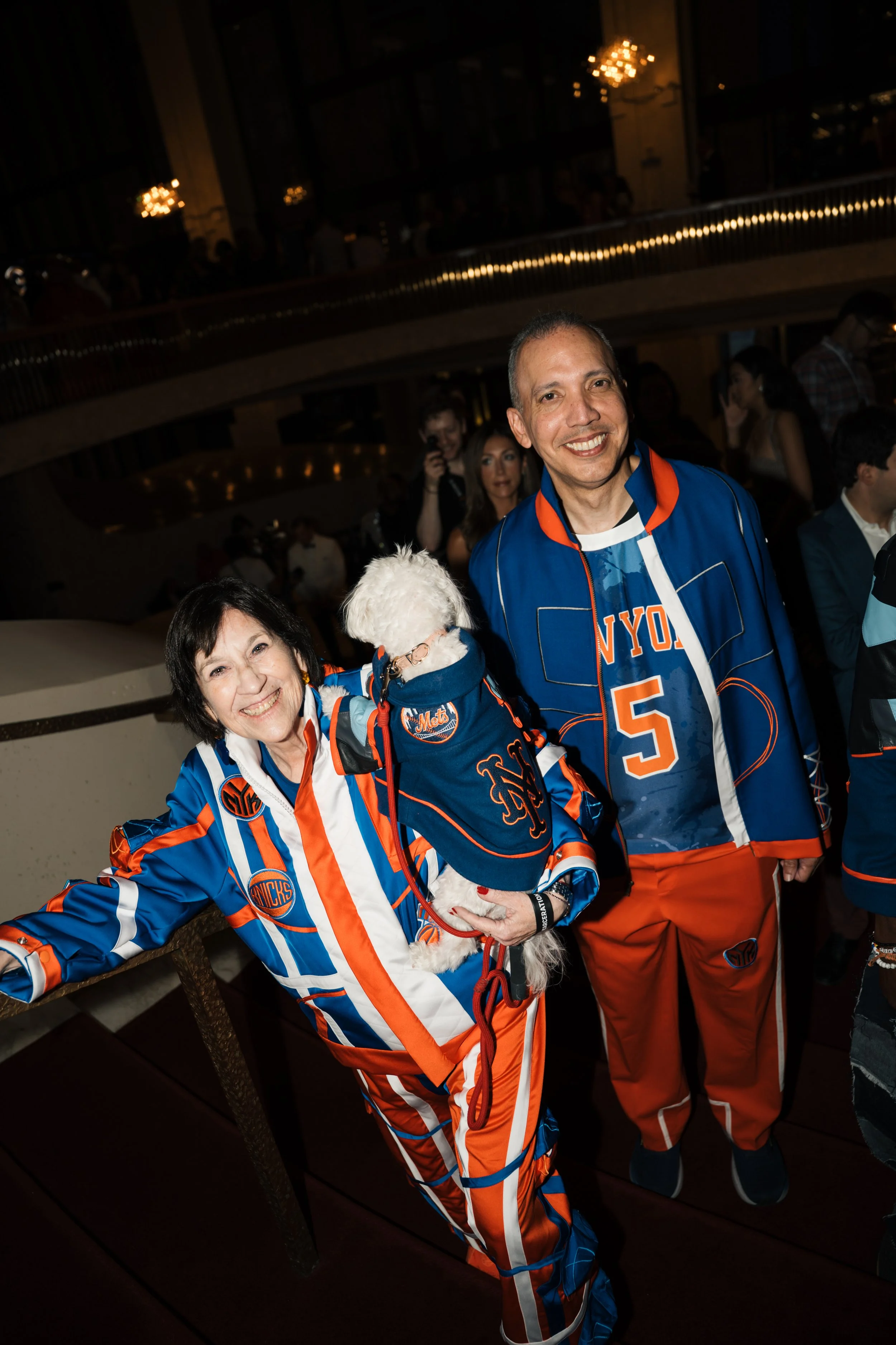 Two people wearing New York Knicks basketball jerseys and a woman holding a small white dog, all smiling at a social event.