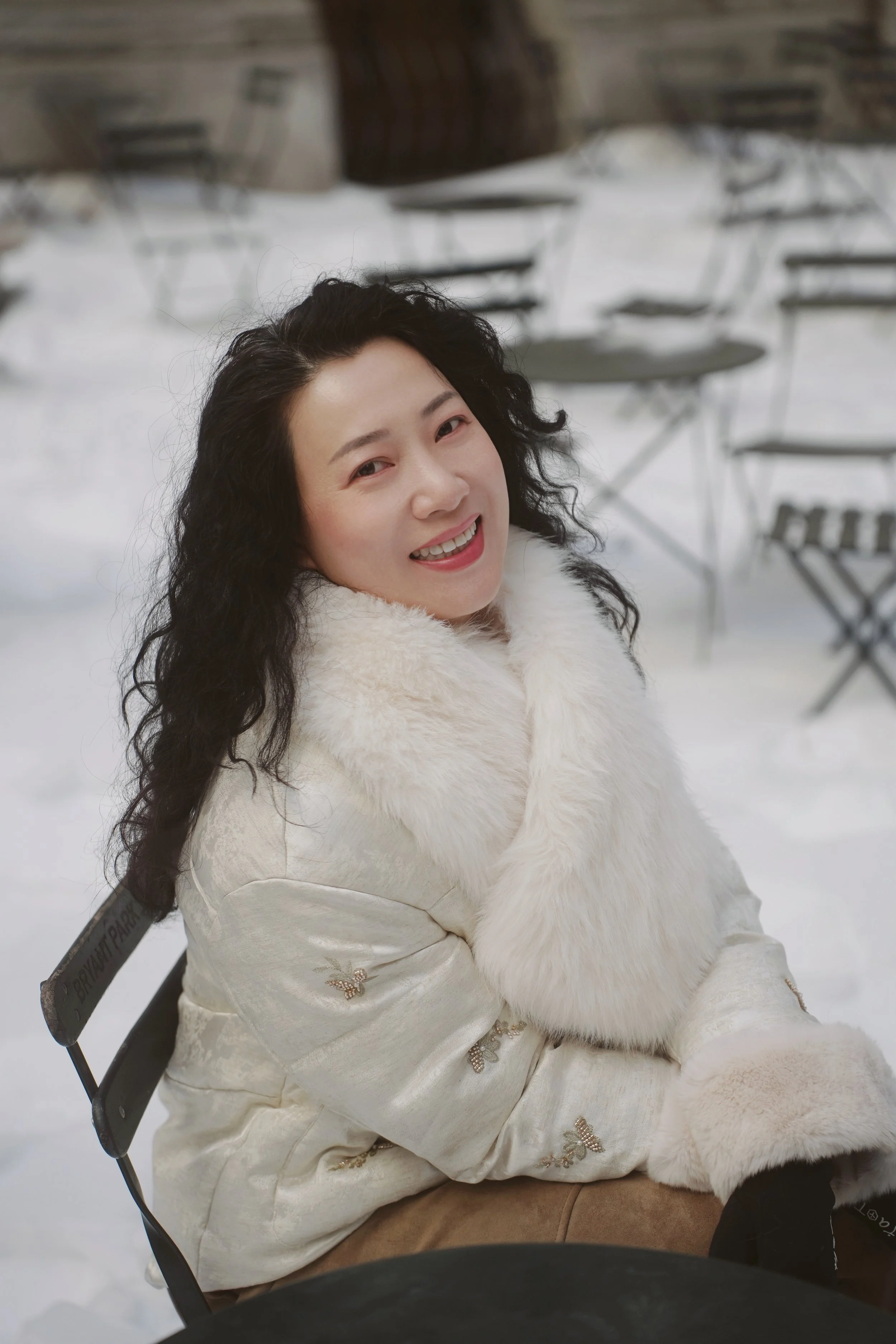 A woman with long dark curly hair smiling and looking up, wearing a white fur coat, sitting outside in a snowy area with empty chairs and tables in the background.