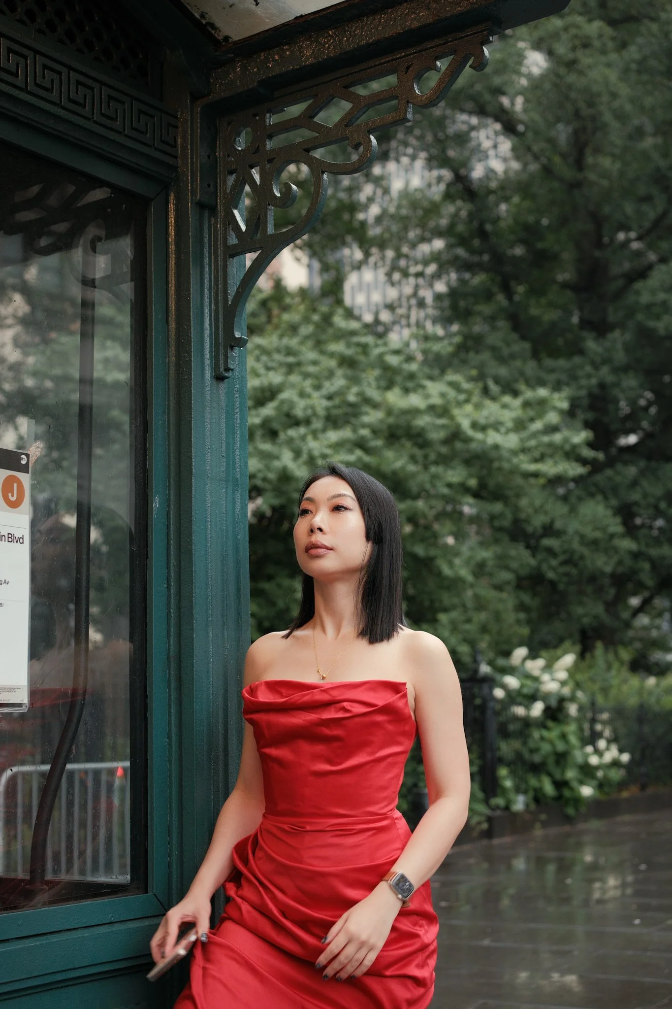 A woman with straight black hair, wearing a strapless red dress, standing outdoors near a green structure with ornate metalwork, and trees in the background.