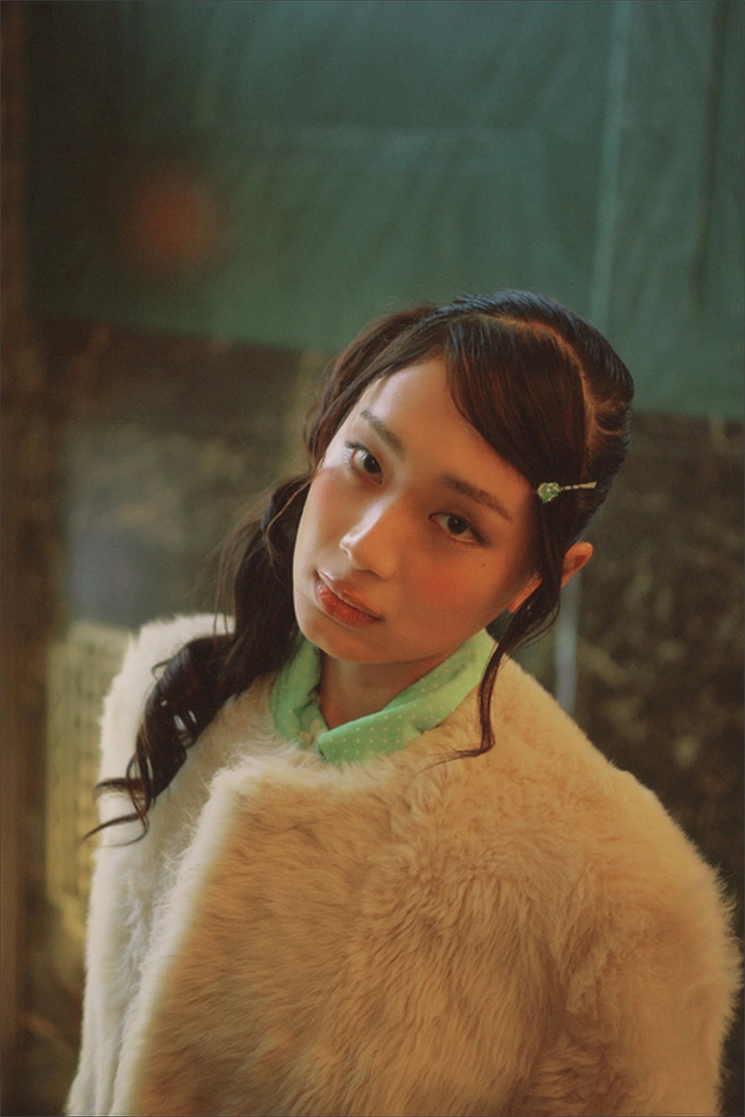 A young woman with dark wavy hair styled with a green hairpin. She is wearing a fur coat and a green polka dot scarf, posing outdoors with blurred trees in the background.
