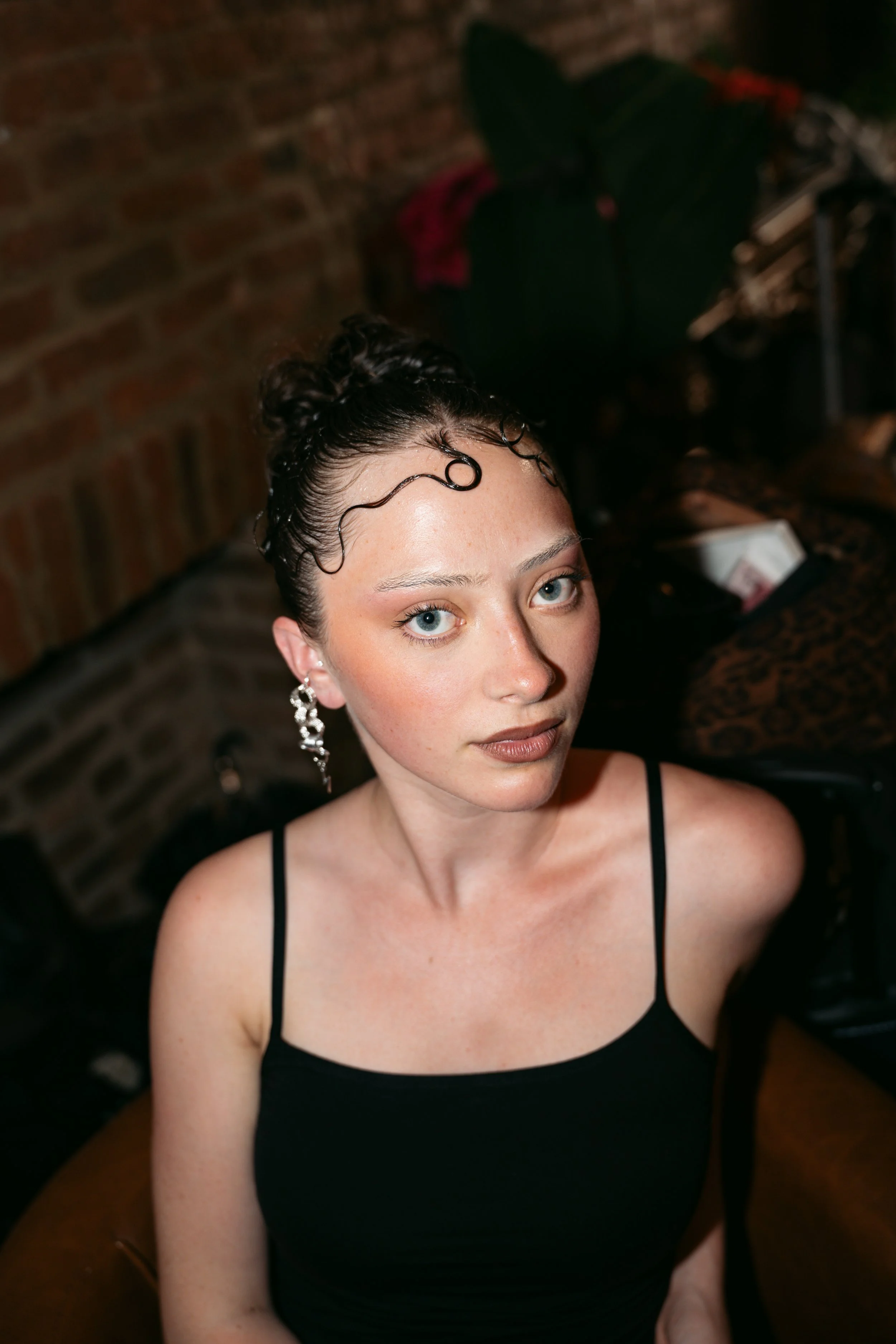 A woman with light skin and blue eyes, wearing a black spaghetti strap dress and ornate earrings, sitting indoors with a brick wall in the background.