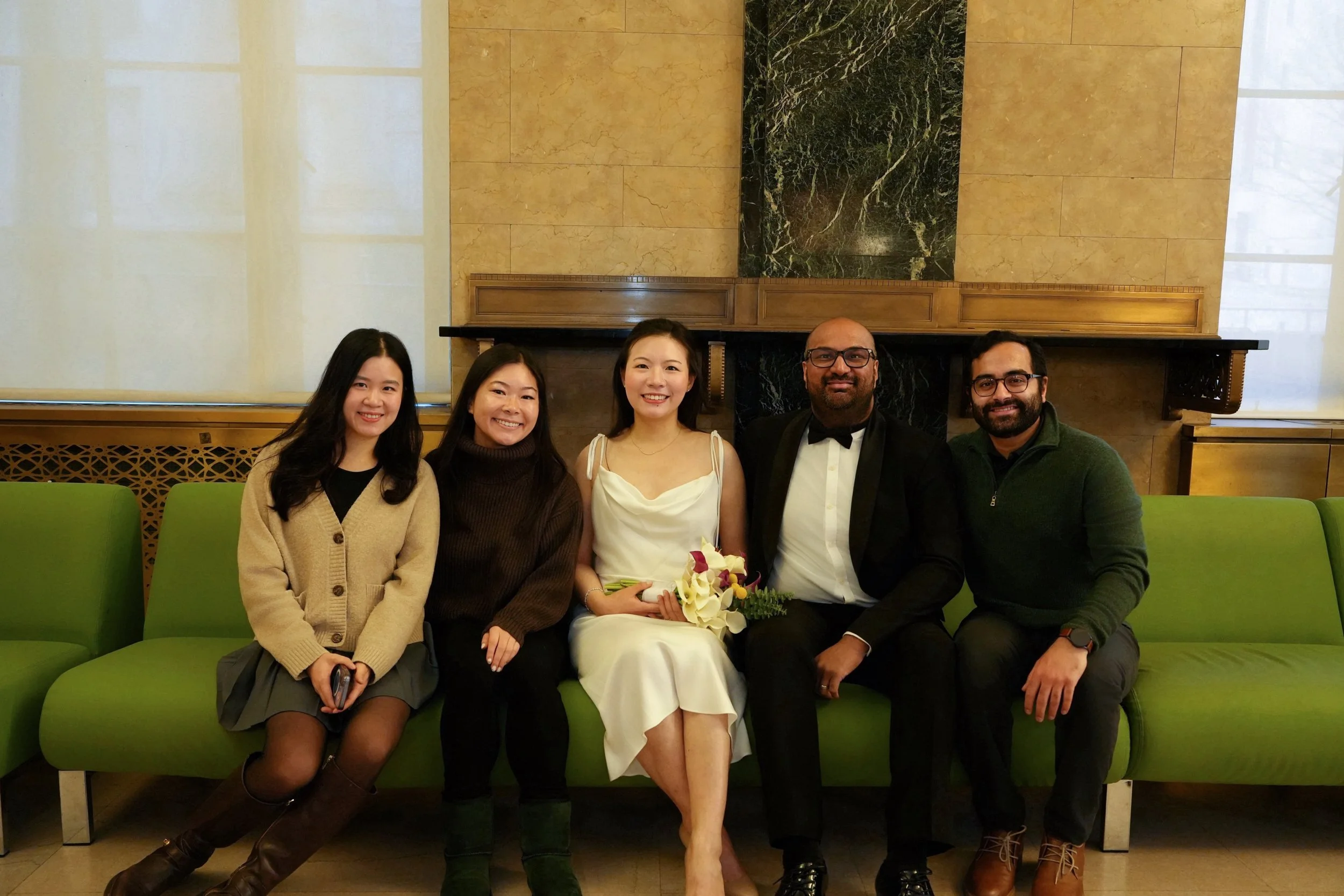 Group of five individuals sitting on a green couch in a room with marble and wooden wall decor, smiling at the camera. The woman in the center wears a white dress and holds a bouquet, suggesting a celebration.