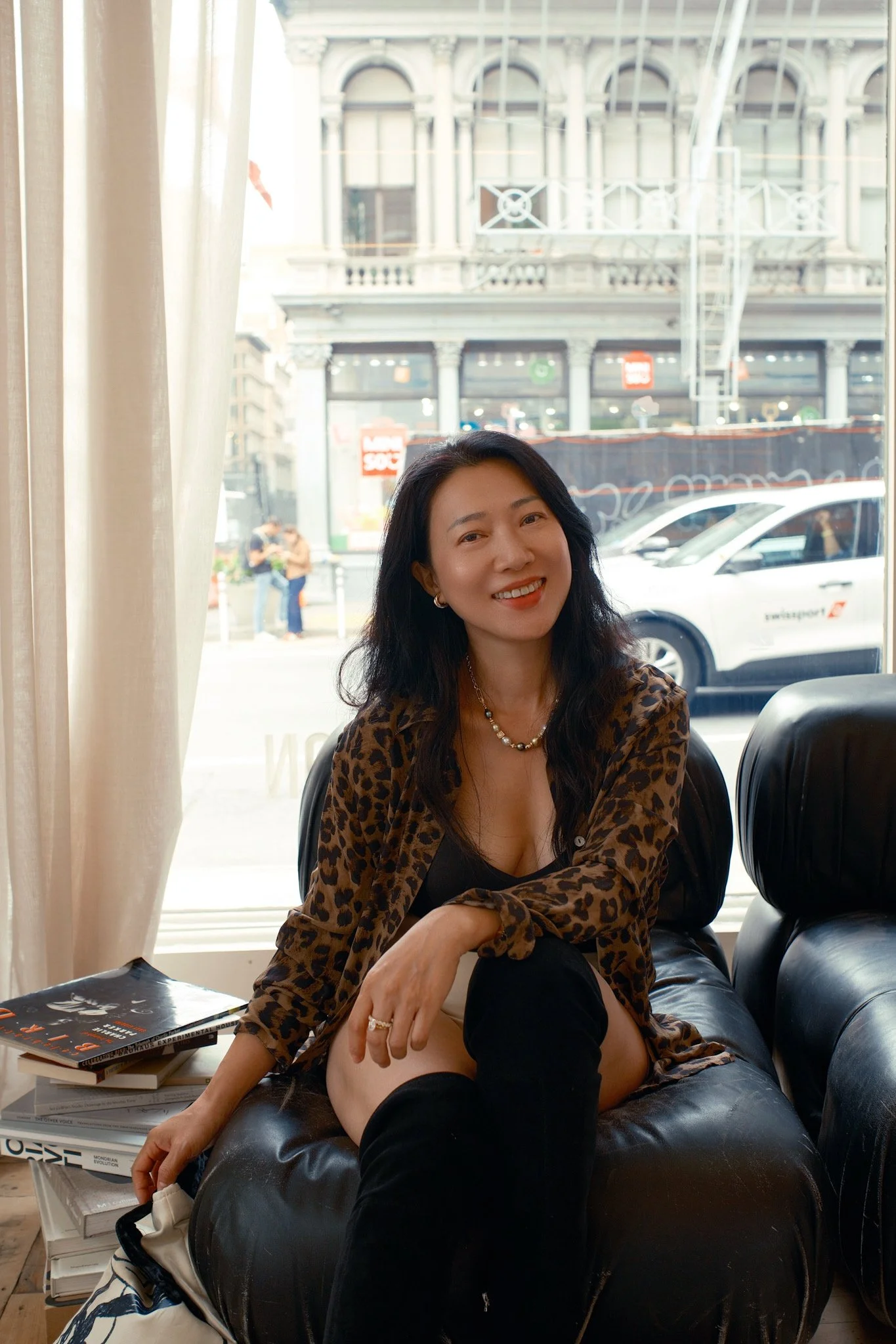 A woman with long black hair, sitting on a black leather couch, smiling, wearing a leopard print jacket, black top, black shorts, and black knee-high socks, with a pearl necklace and rings, in a cafe with large windows showing a city street with hist