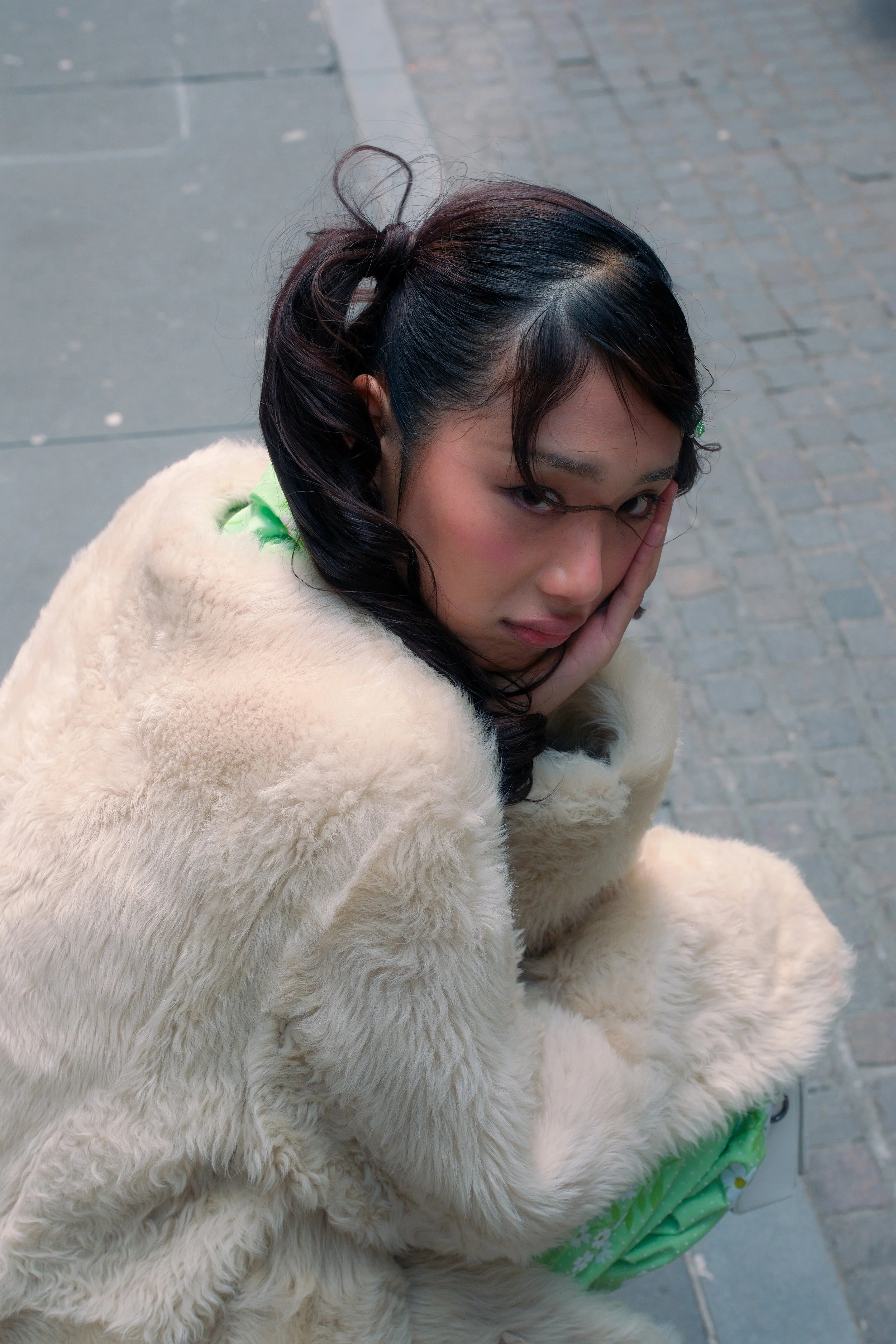 A person with dark hair styled in a high ponytail with loose bangs, resting her face on her hand, wearing a fluffy cream-colored coat.