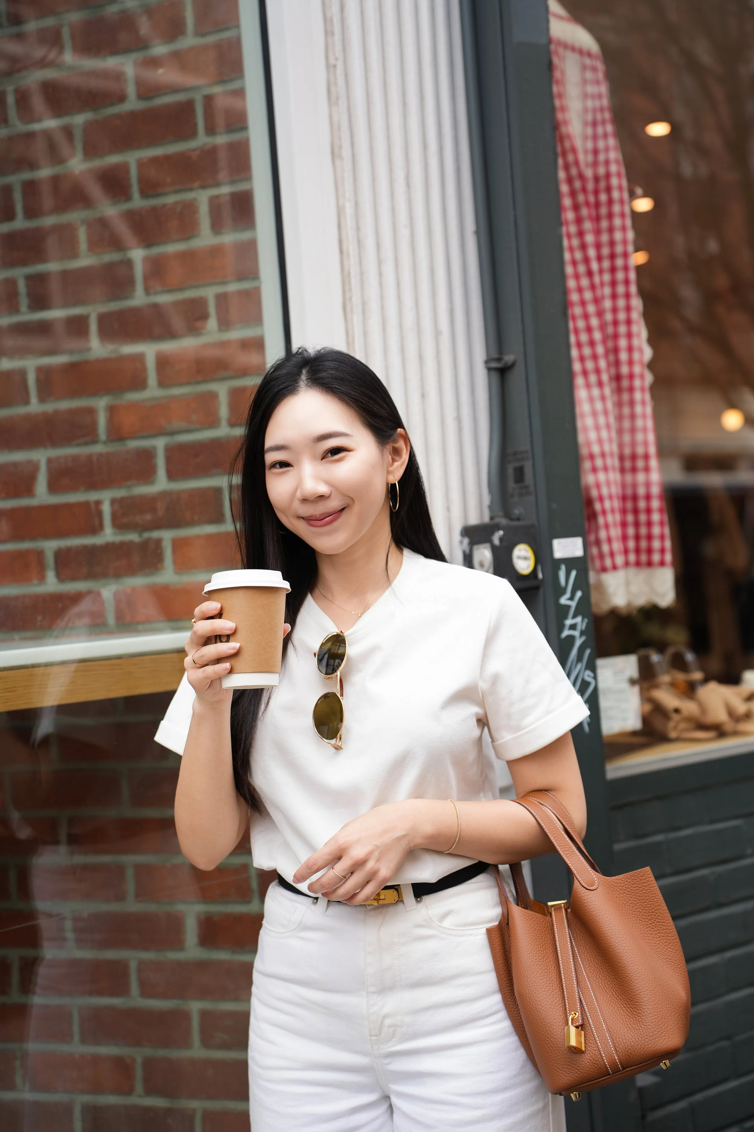 A young woman with long dark hair holding a disposable coffee cup, standing outdoors in front of a brick wall and a storefront, wearing a white t-shirt, white pants, sunglasses hanging from her shirt, gold jewelry, and carrying a brown handbag.