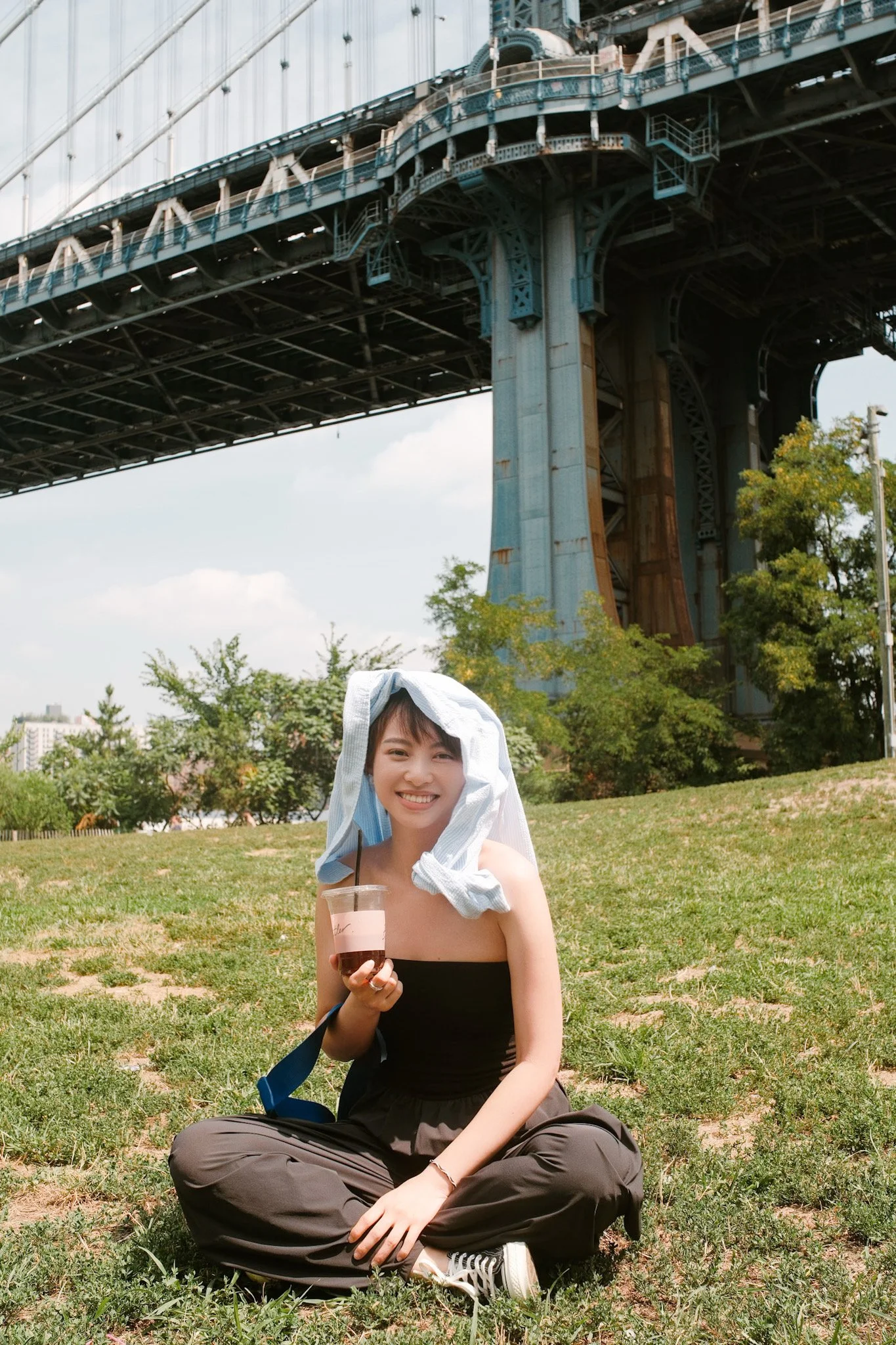 A smiling woman sitting cross-legged on grass in a park, holding a drink and wearing a sun hat with a towel draped over her head, with a bridge and trees in the background.