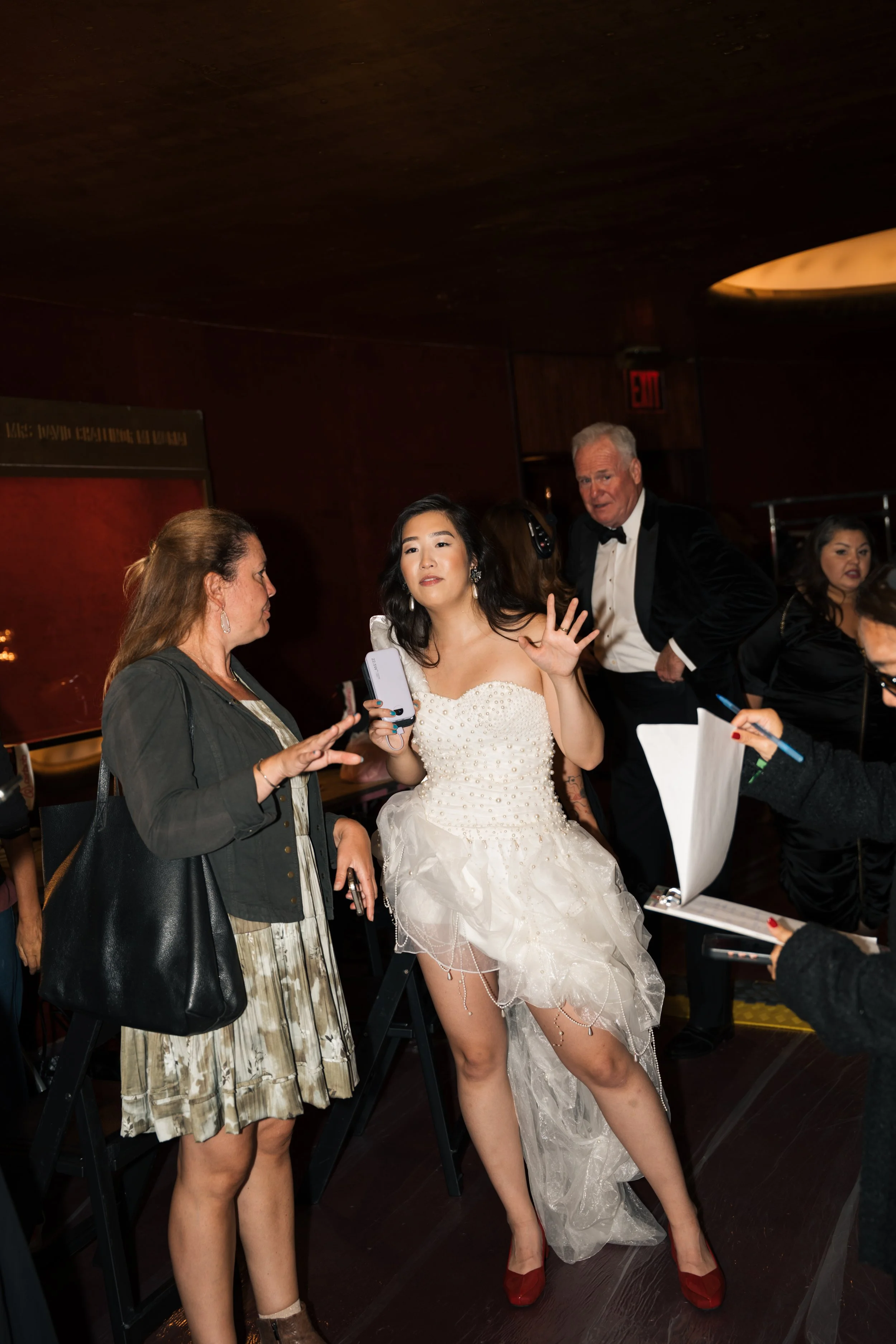 A woman in a wedding dress with a sheer overlay and red shoes poses with her hand raised. She is surrounded by other women and a man in a tuxedo in a dimly lit indoor setting, possibly backstage or at a wedding reception.