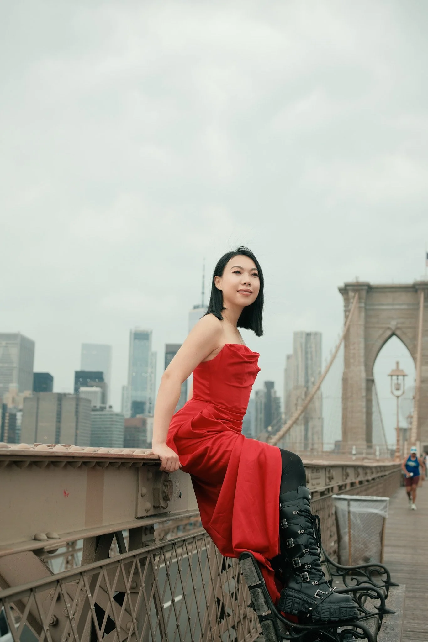 A woman in a red dress sitting on a park bench on the Brooklyn Bridge with New York City skyline in the background.