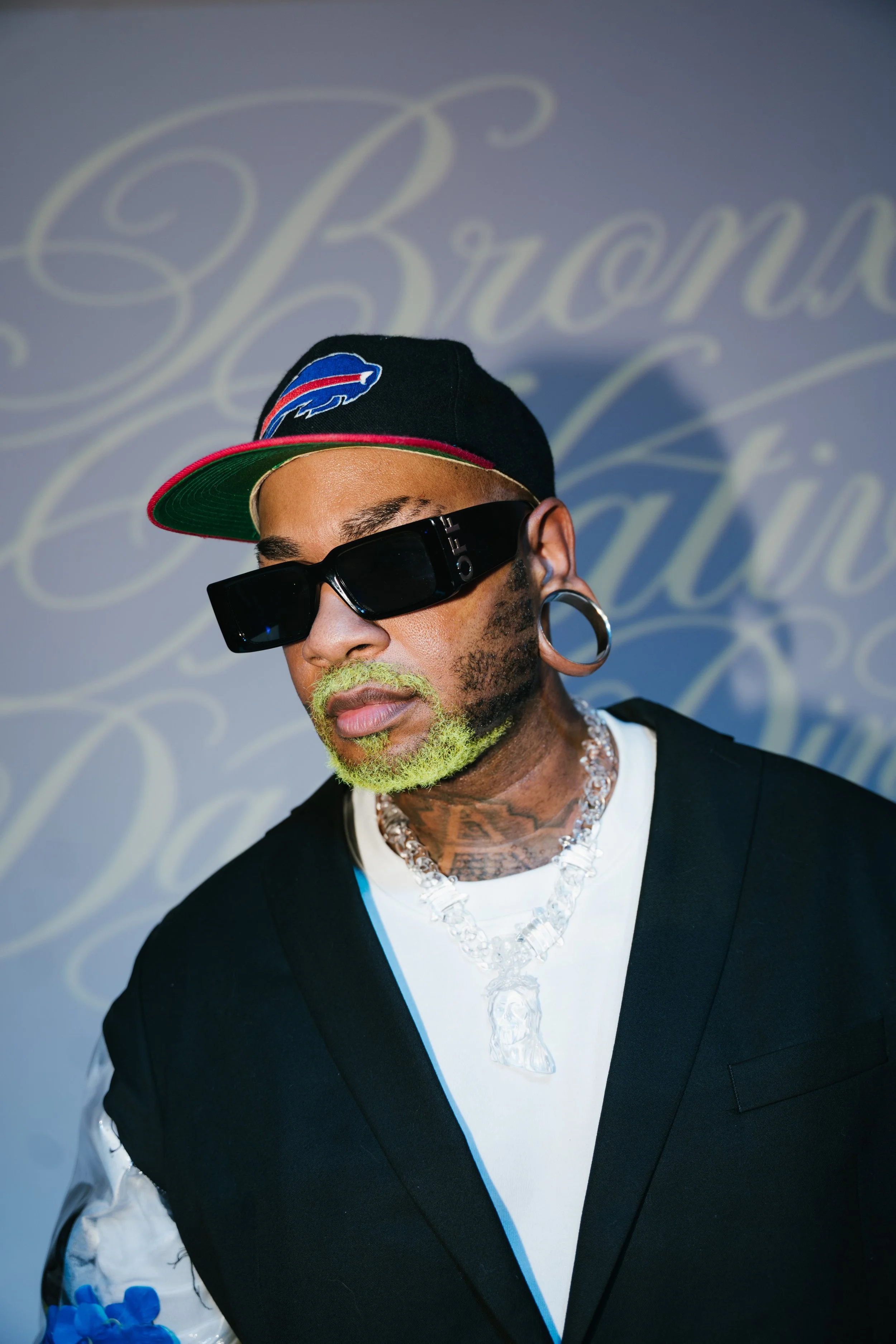 A man wearing a black baseball cap with the Buffalo Bills logo, dark sunglasses, earrings, and green facial hair, dressed in a black blazer over a white shirt with a clear necklace, standing in front of a background with white cursive writing.
