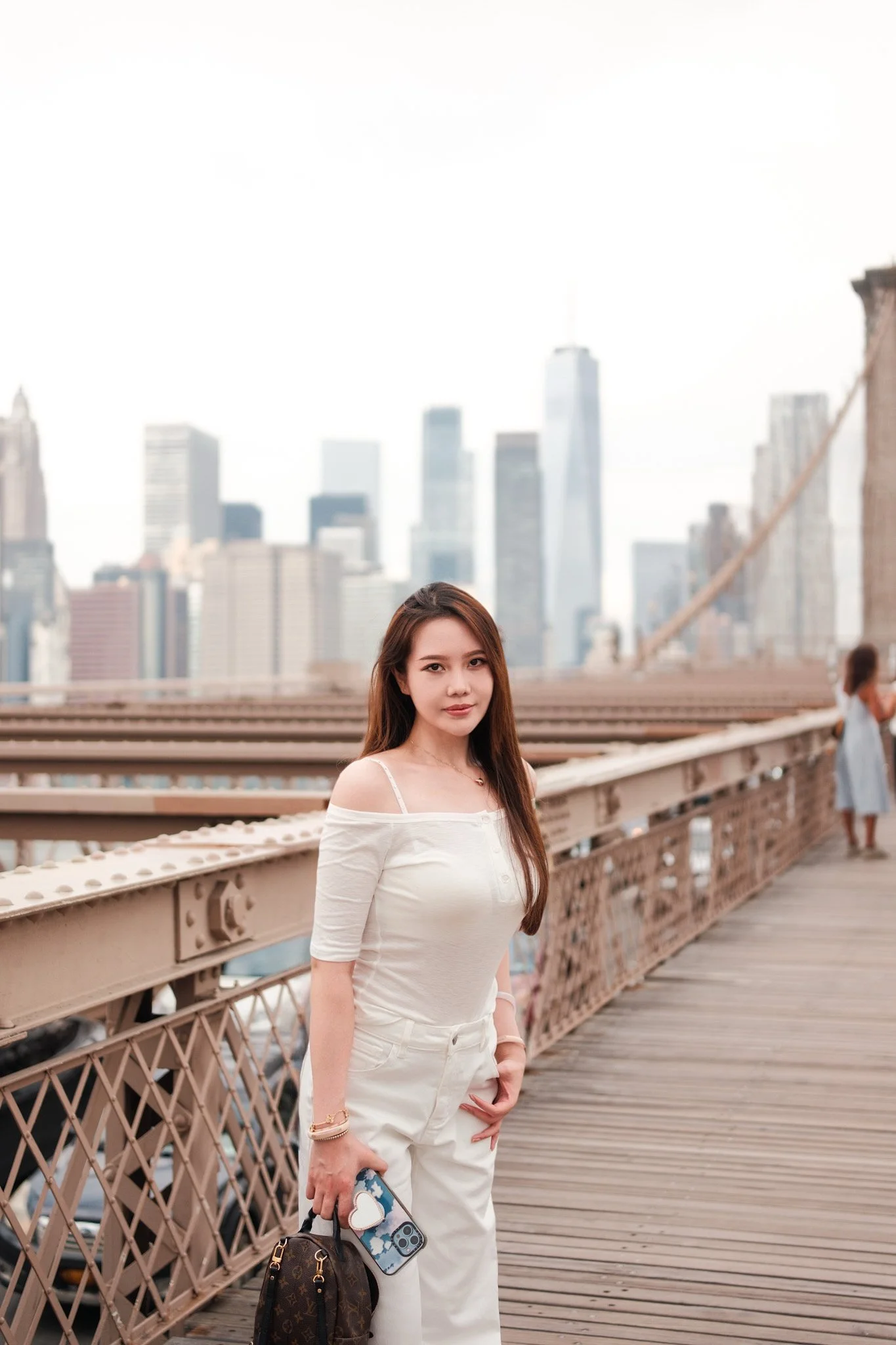 A young woman with long brown hair standing on the Brooklyn Bridge in New York City, holding a Louis Vuitton backpack and a smartphone with a heart-shaped pop socket, with the city skyline in the background.