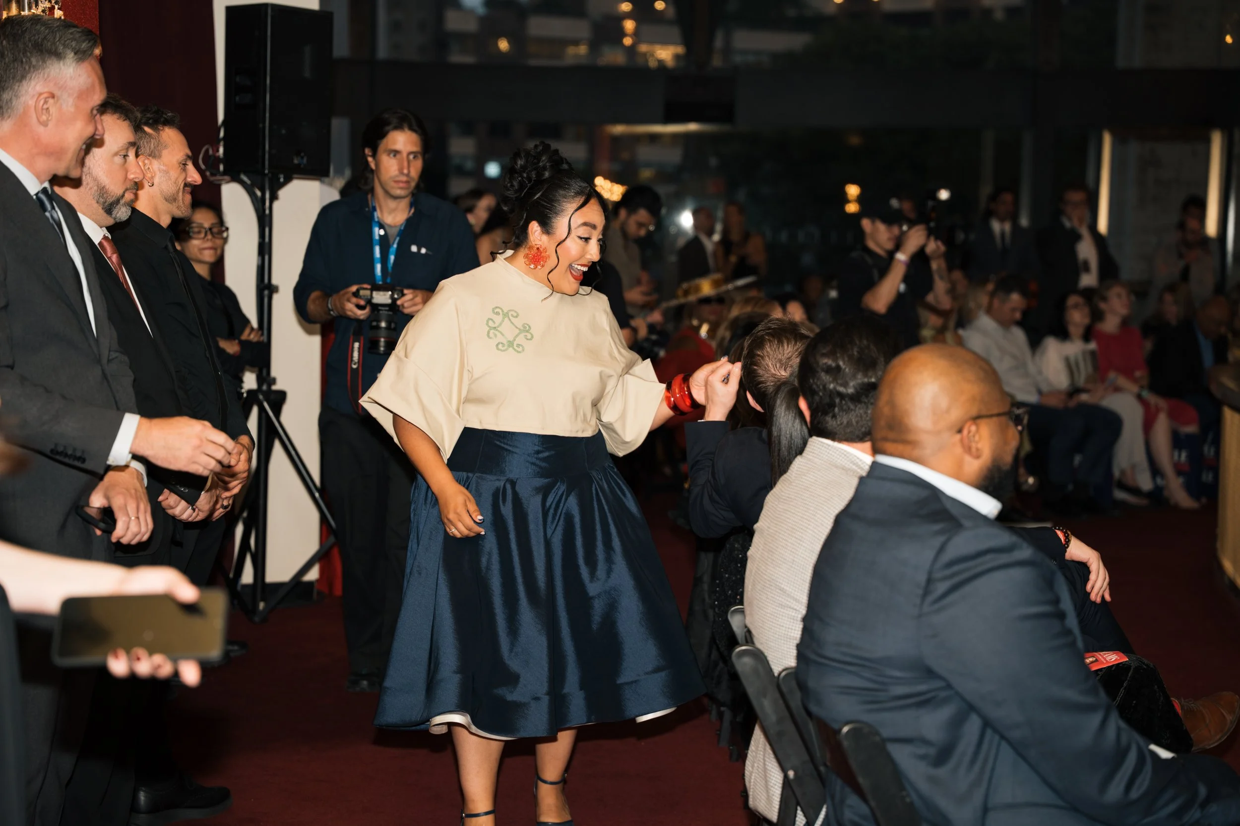 A woman with dark hair styled in a bun, wearing a cream top with green embroidery and a long navy skirt, smiling and interacting with a seated man at an indoor event. Other people are standing and sitting, watching or taking photos, in a lively, form