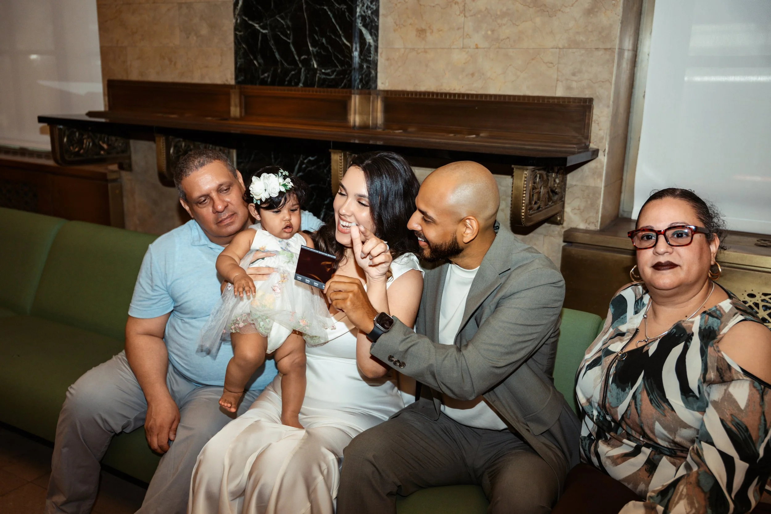Group of five people sitting on a green bench in a room with marble walls. A woman in a white dress is holding a little girl with a floral headband, who is looking at a card held by a man in a gray suit. The man is smiling and pointing at the card. T