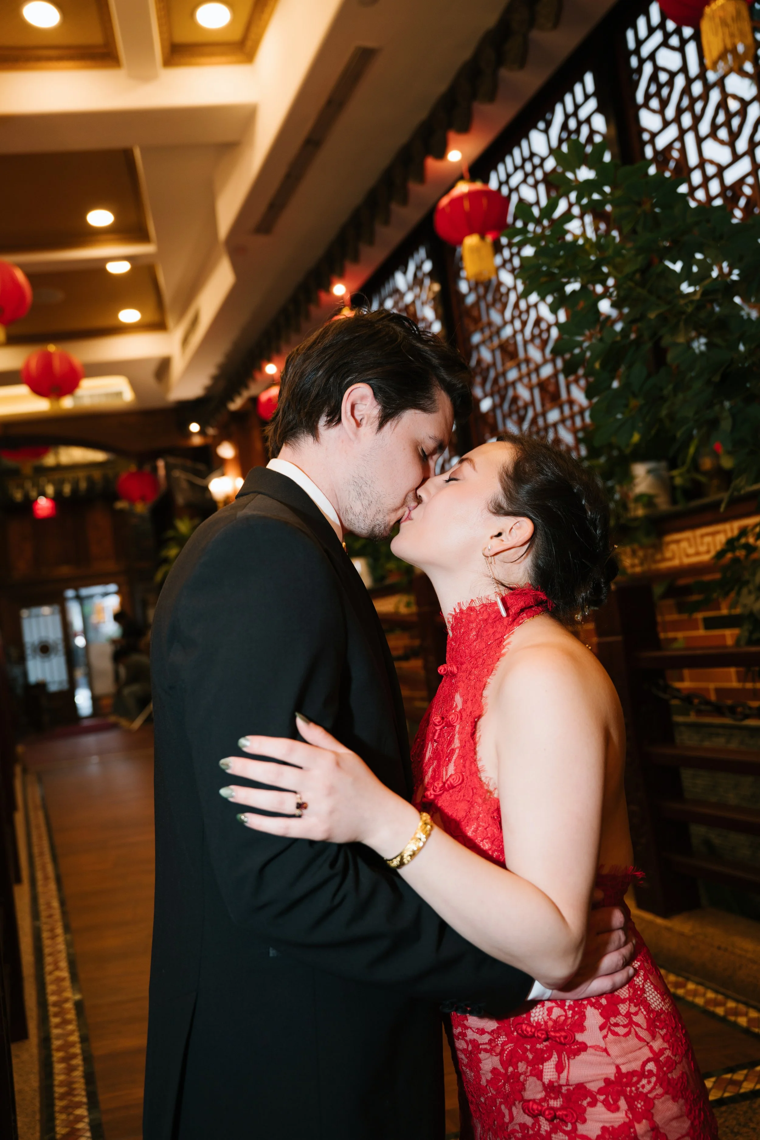 A couple, a man in a black suit and a woman in a red lace dress, kissing each other in an indoor setting decorated with red Chinese lanterns and wooden lattice panels.