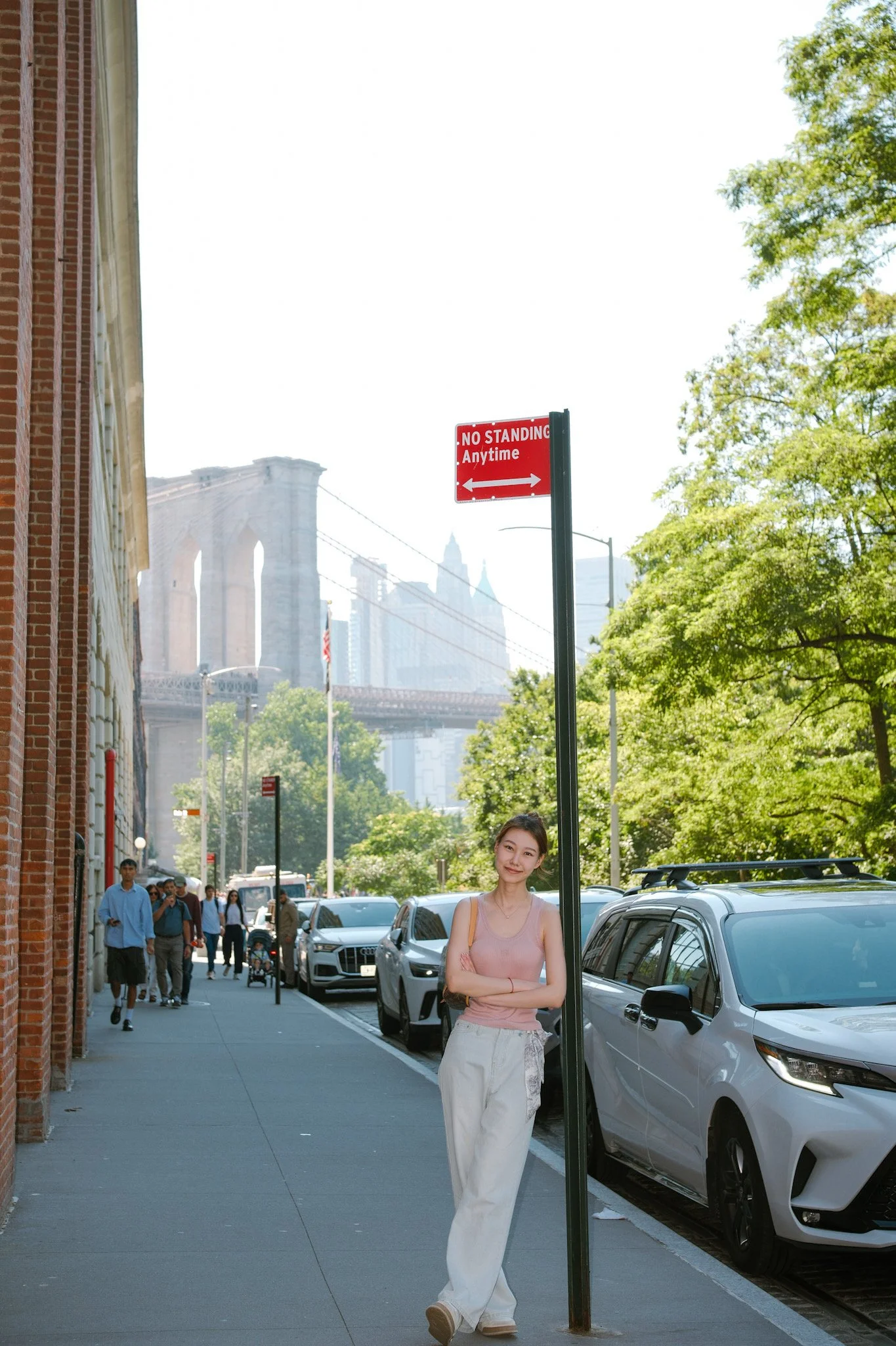 Young woman stands on city sidewalk with arms crossed, smiling near a parking meter under a street sign that says 'No Standing Anytime,' with the Brooklyn Bridge in the background.
