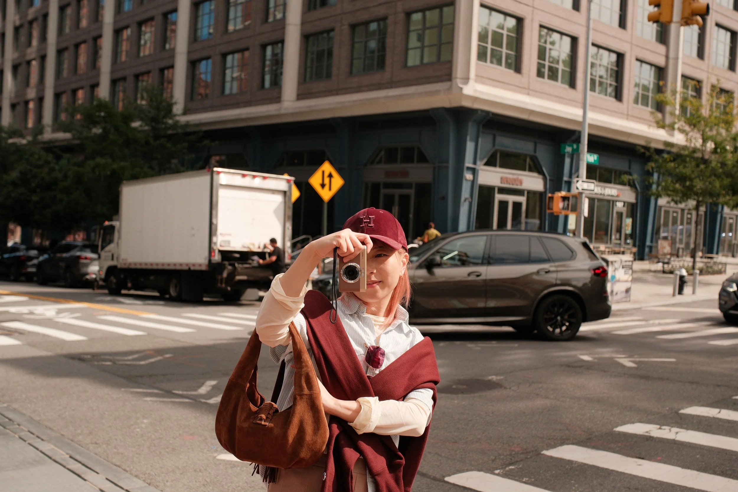 A young woman with red hair taking a photo with a vintage camera at a city crosswalk, with cars and a large building in the background.