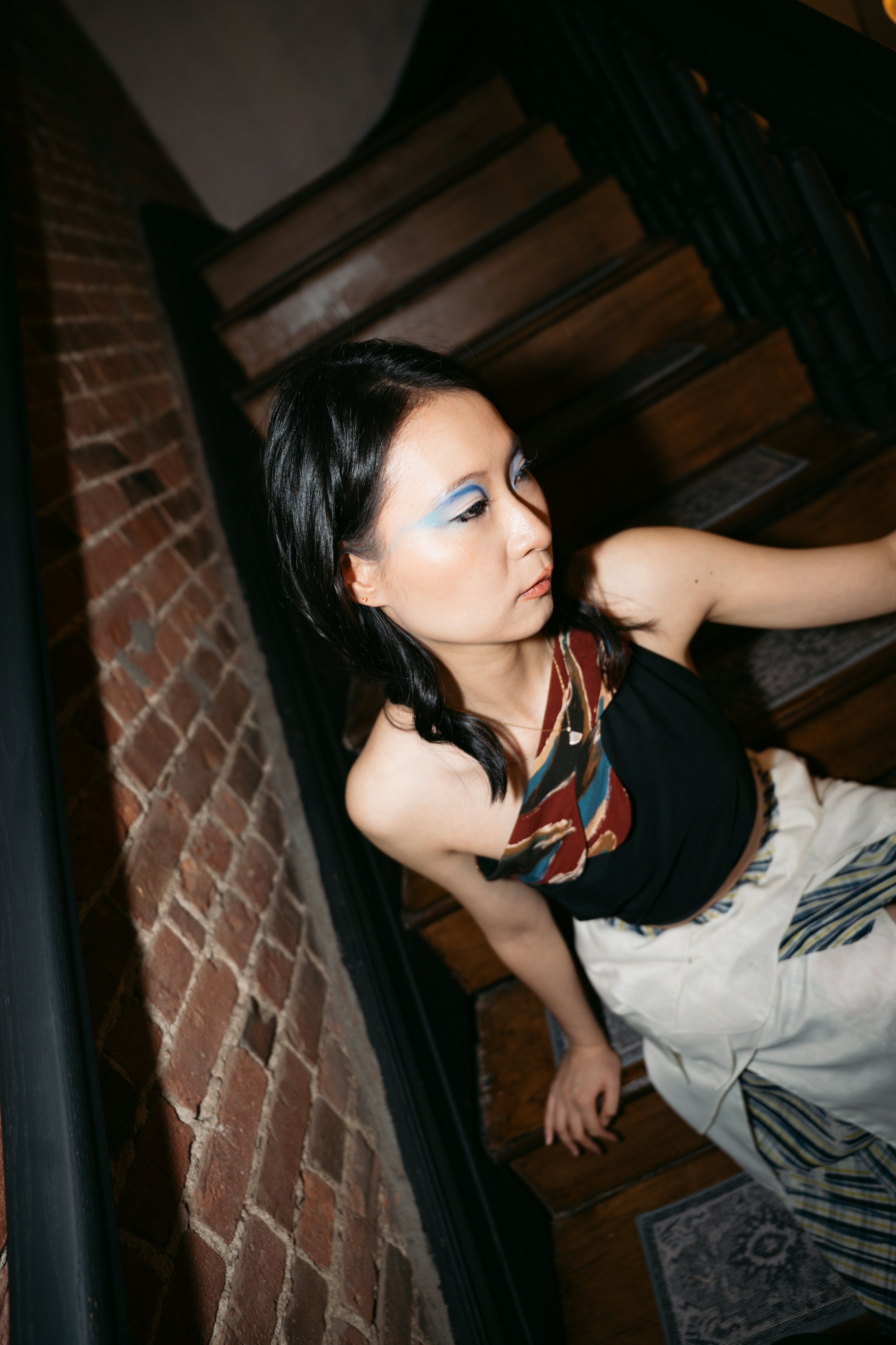 Young woman with dark hair and blue eyeshadow sitting on wooden stairs next to a brick wall, wearing a colorful patterned top and light-colored pants.