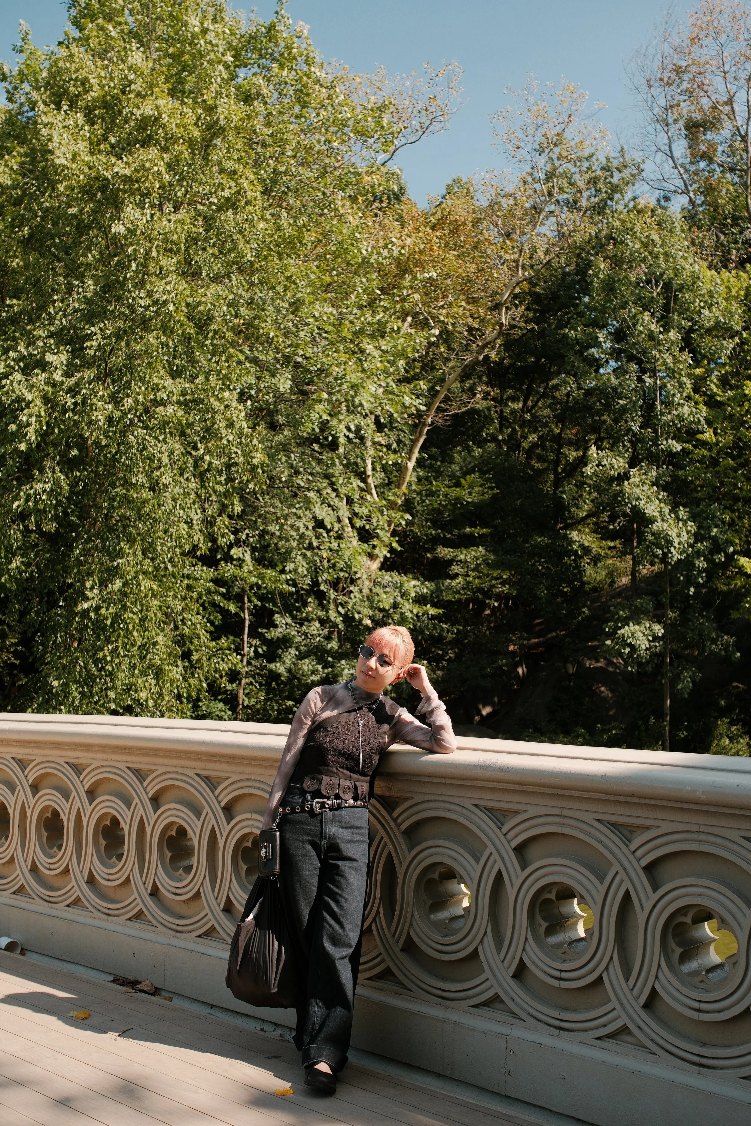 A woman with short hair and sunglasses standing and leaning against a decorative bridge railing in a park with trees and blue sky in the background.