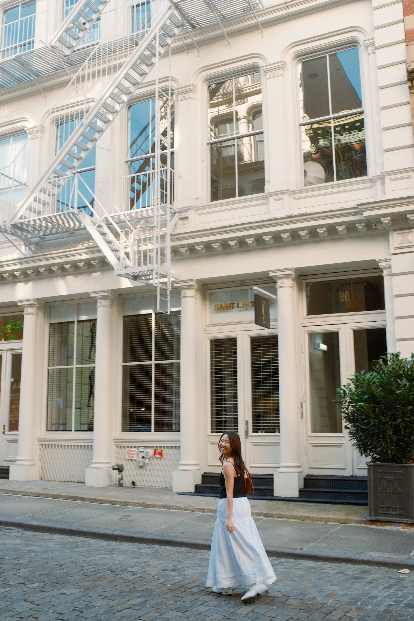 A woman wearing a black top and white skirt walking down a city street with a large white building featuring tall windows and fire escape stairs behind her, and a small tree to the right.