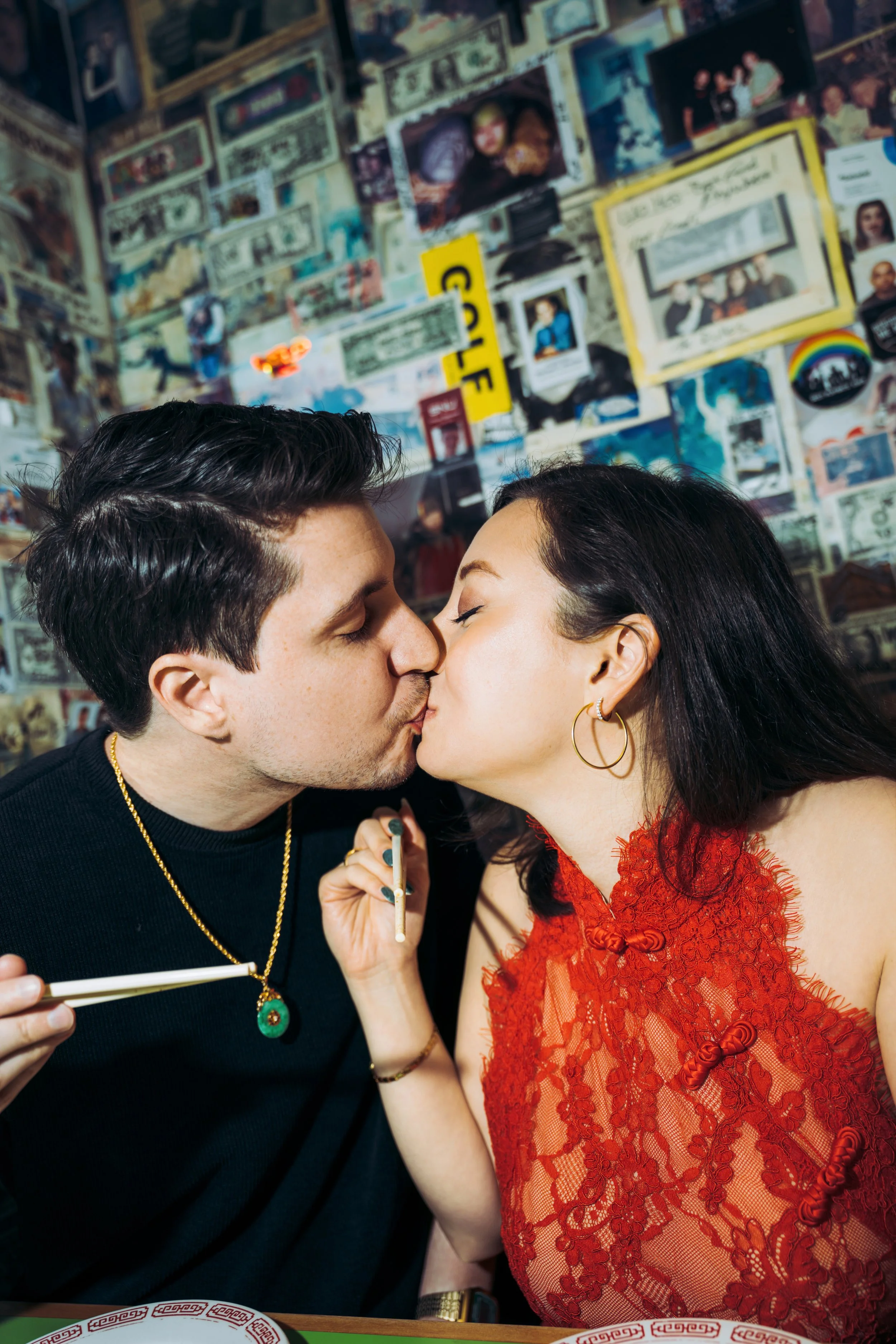 A young man and woman are sharing a kiss in a restaurant or cafe. The man has dark, wavy hair and is wearing a black shirt with gold jewelry, including a chain and pendant. The woman has dark hair and is wearing a red lace dress with floral patterns 
