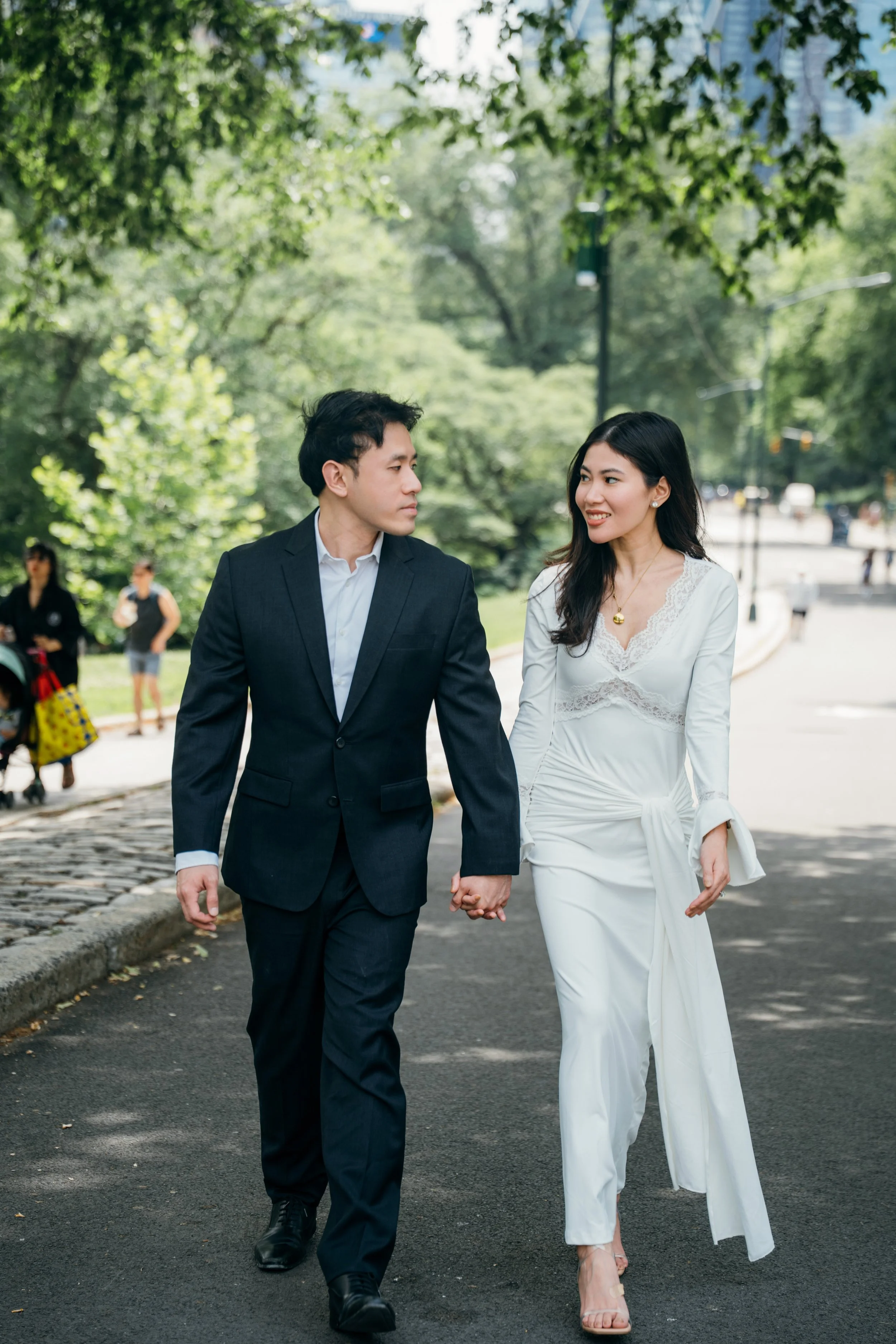 A couple, a man and a woman, holding hands and walking outdoors on a paved path in a park with lush green trees around them. The woman is wearing a white outfit with a lace detail, and the man is in a dark suit.