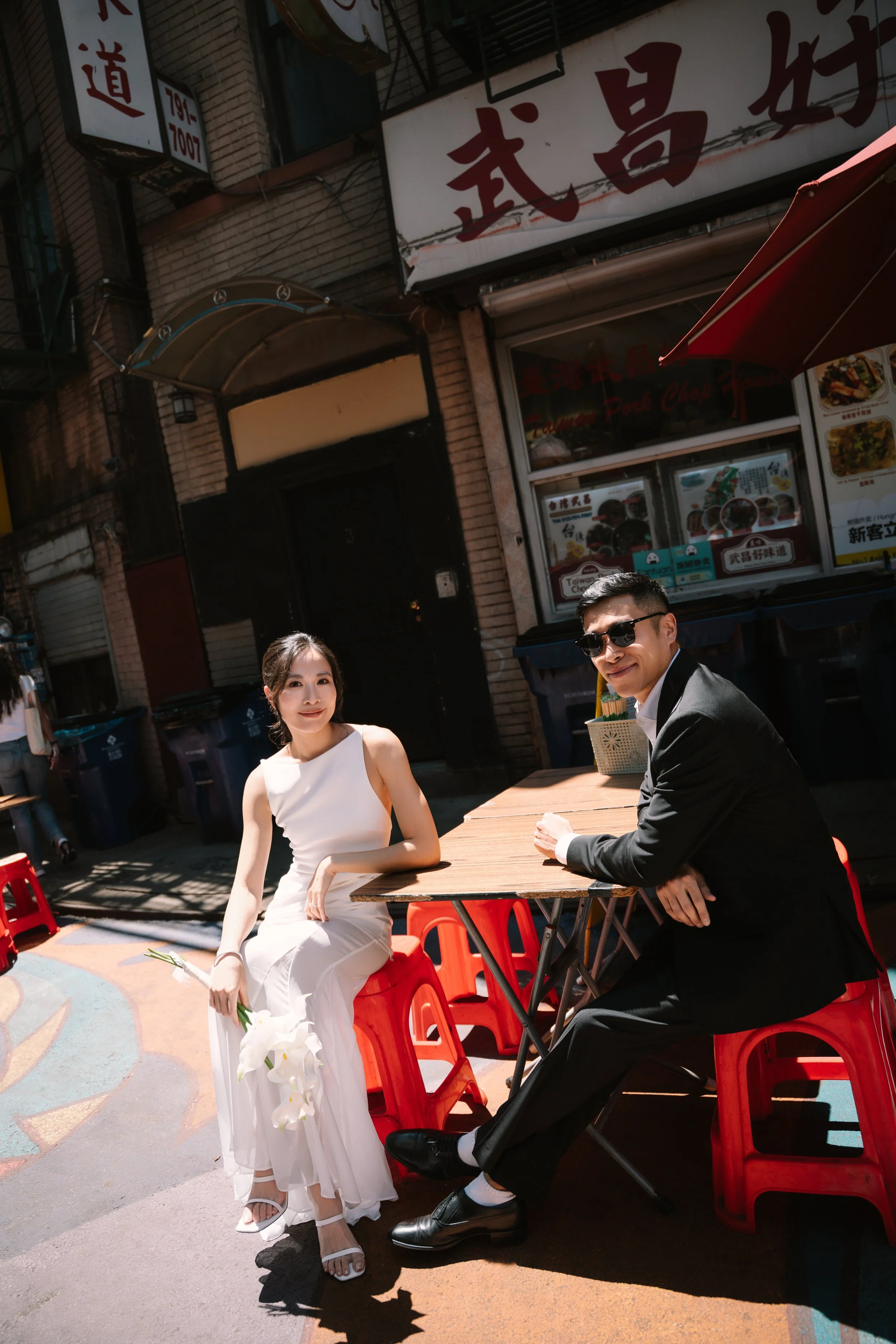 A man and woman sit outdoors at a street-side table, with the woman dressed in a white gown holding flowers, and the man in a black suit with sunglasses. Behind them is a food stand with signs in Chinese characters, and red plastic stools.