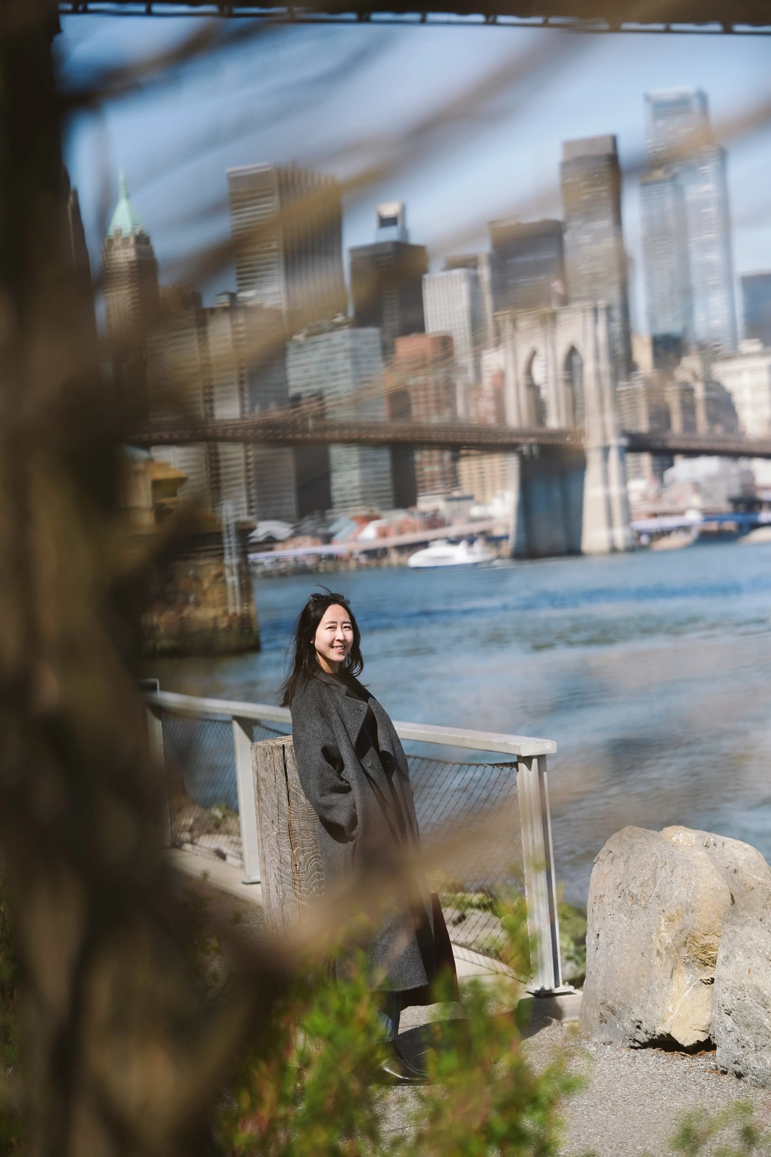 A woman with long black hair smiling, wearing a gray coat, standing by a waterfront with the New York City skyline including the Brooklyn Bridge and skyscrapers in the background, seen through a blurred foreground of foliage.