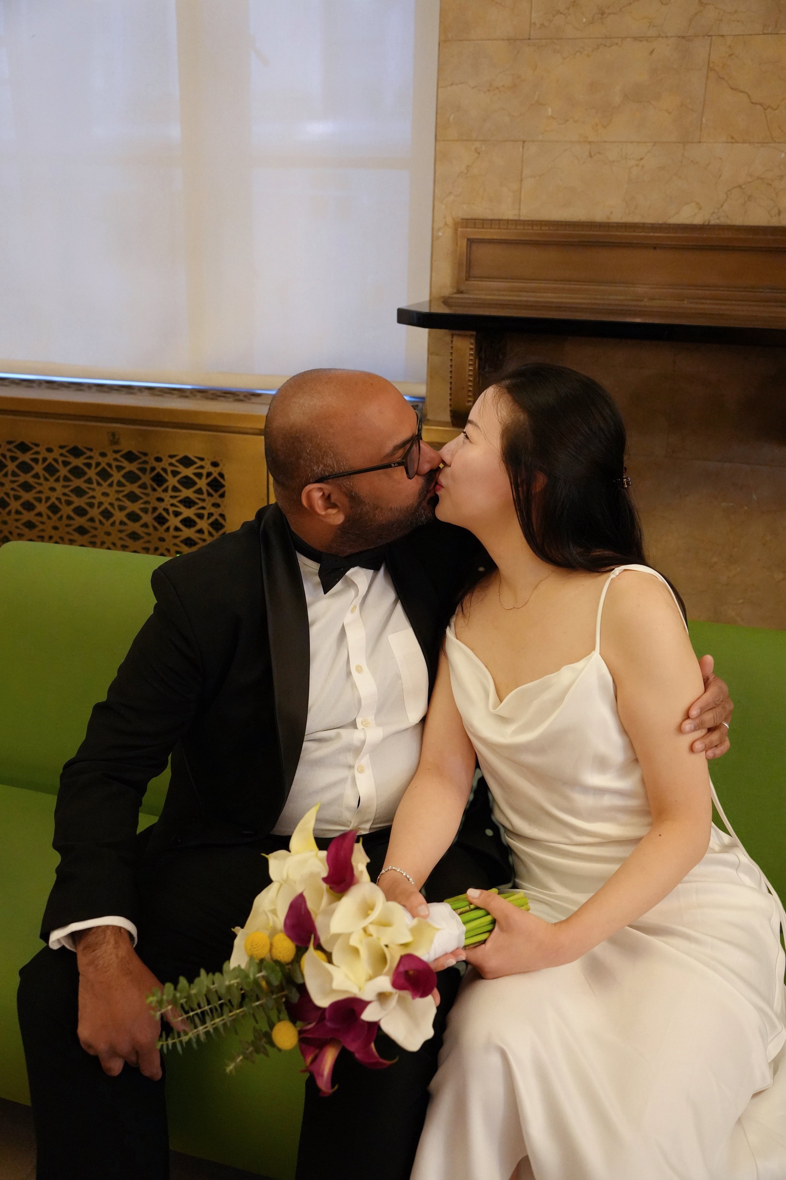 A couple dressed in formal attire, the man in a tuxedo and the woman in a wedding dress, sharing a kiss. The woman is holding a bouquet of flowers, and they are sitting on a green sofa in a room with a marble wall and wood paneling.