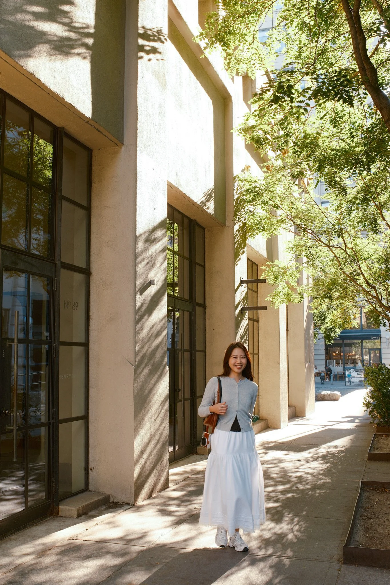 A young woman with long hair smiling and walking along a sidewalk with trees and modern buildings in the background, sunlight casting shadows on the wall.