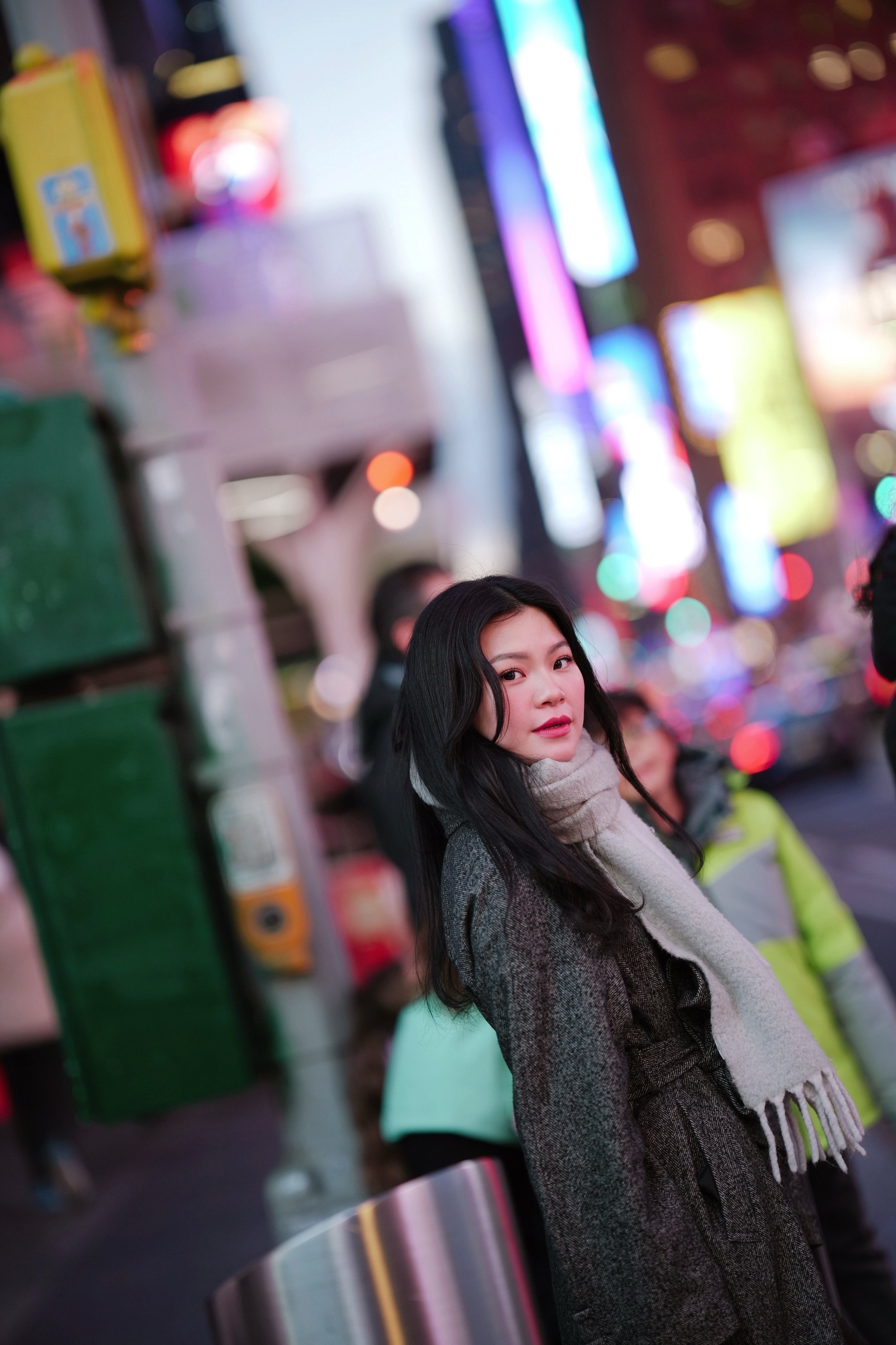 A young woman with dark hair in a gray coat and scarf standing at a subway station with colorful, illuminated advertisements in the background.