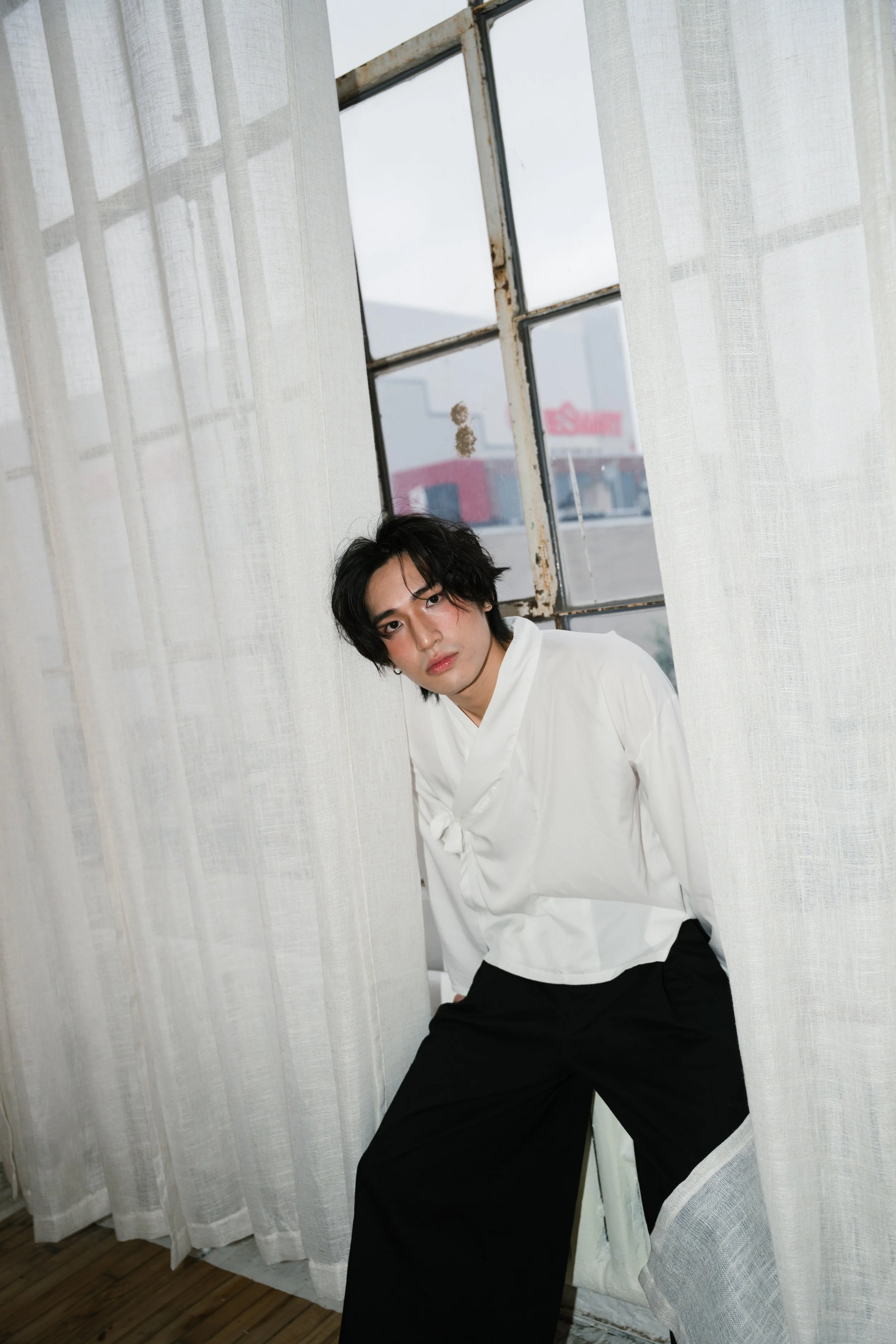 A young man with dark, wavy hair and light skin standing indoors near a window with white curtains, wearing a white long-sleeved shirt and black pants, looking directly at the camera.
