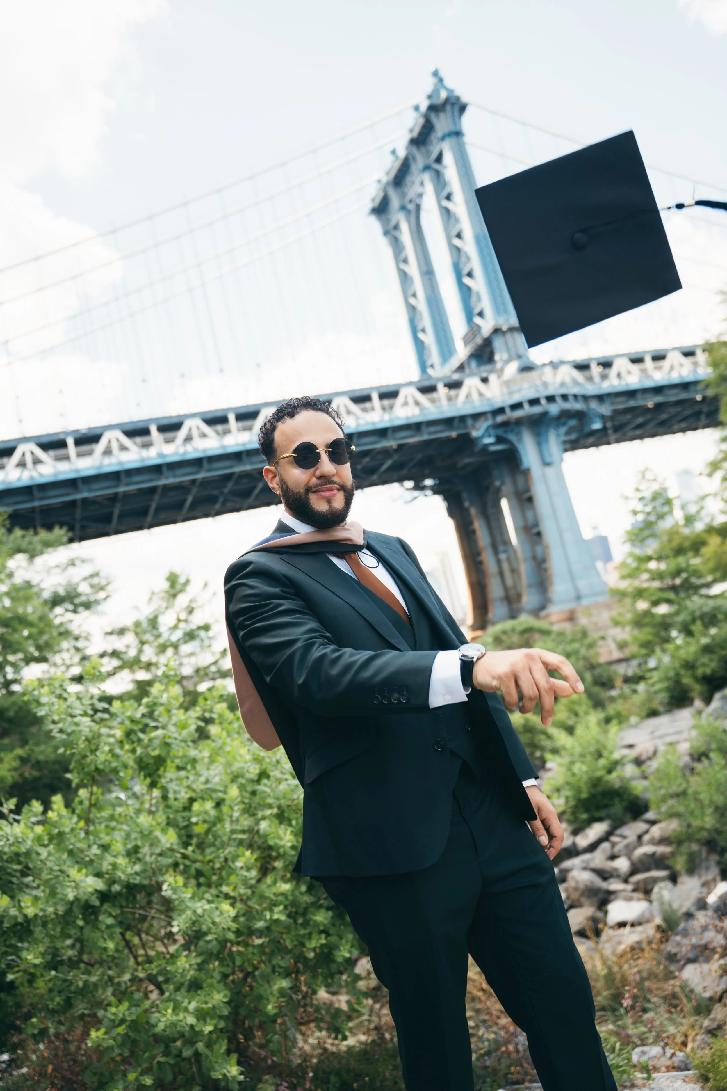 A man in a tuxedo with sunglasses and a graduation stole standing outdoors under a bridge, with green foliage and rocks around, during daytime.