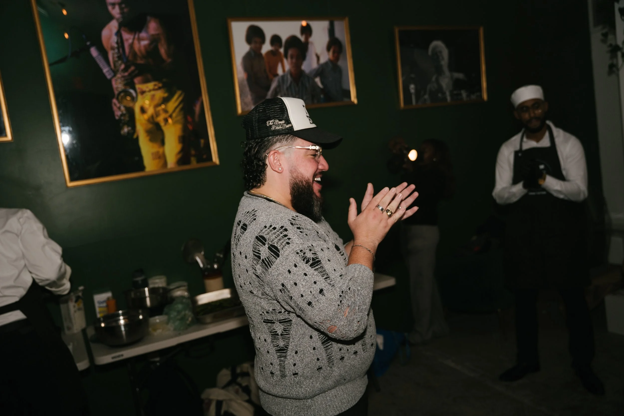 A man with curly hair and a beard, wearing a gray sweater with black patterns and a black Trucker hat, is clapping and smiling in a room with framed photos on the green wall behind him. There is a person in a white chef uniform and glasses standing t